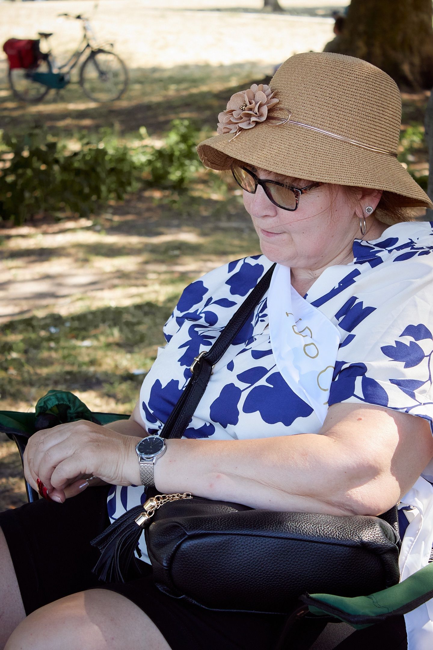 Elderly woman in straw hat with flower, glasses, and floral shirt sitting in a park, looking down at her bag.
