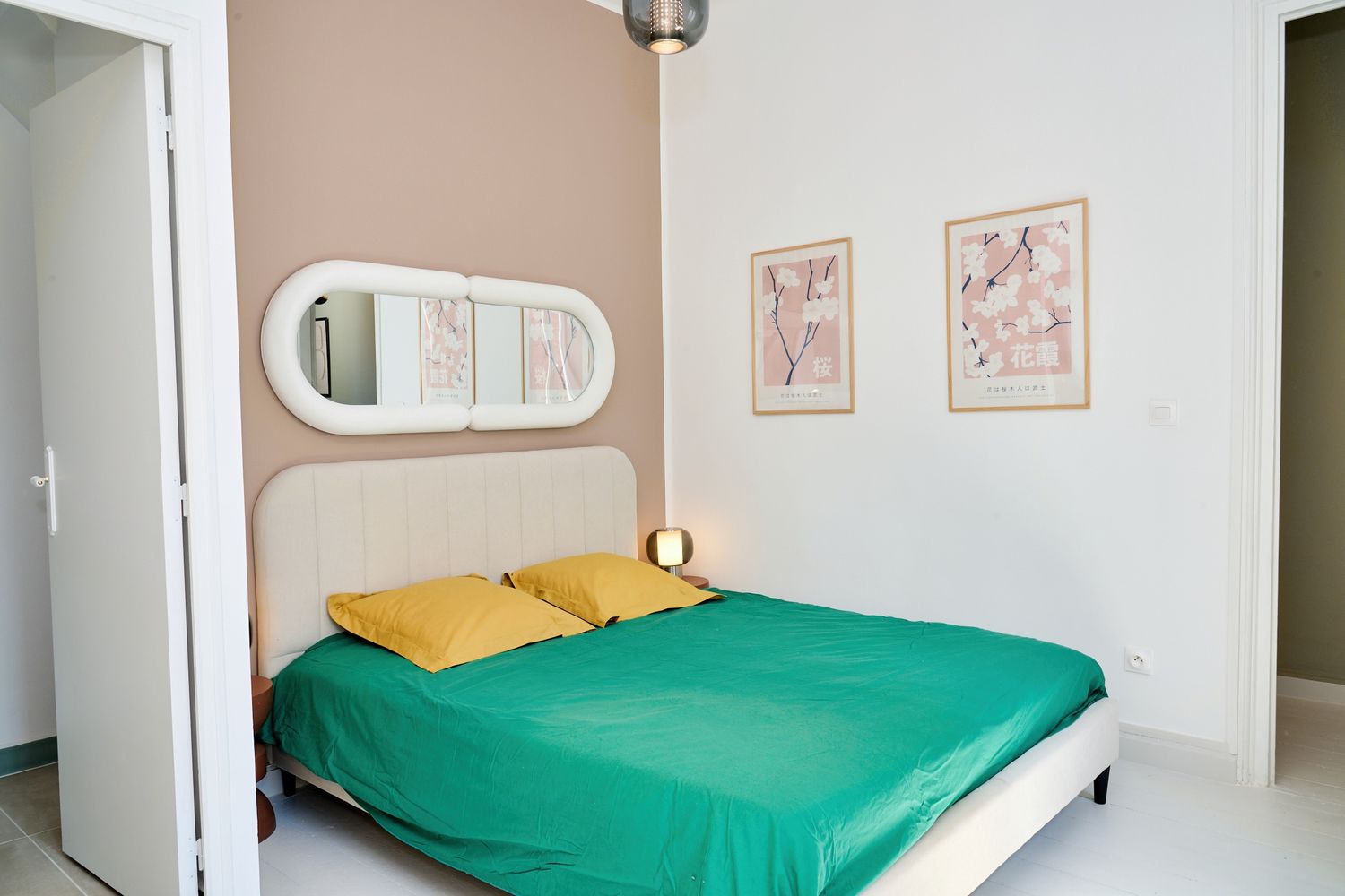 Bright bedroom with green bedspread, yellow pillows, oval mirror, and pastel framed wall art.