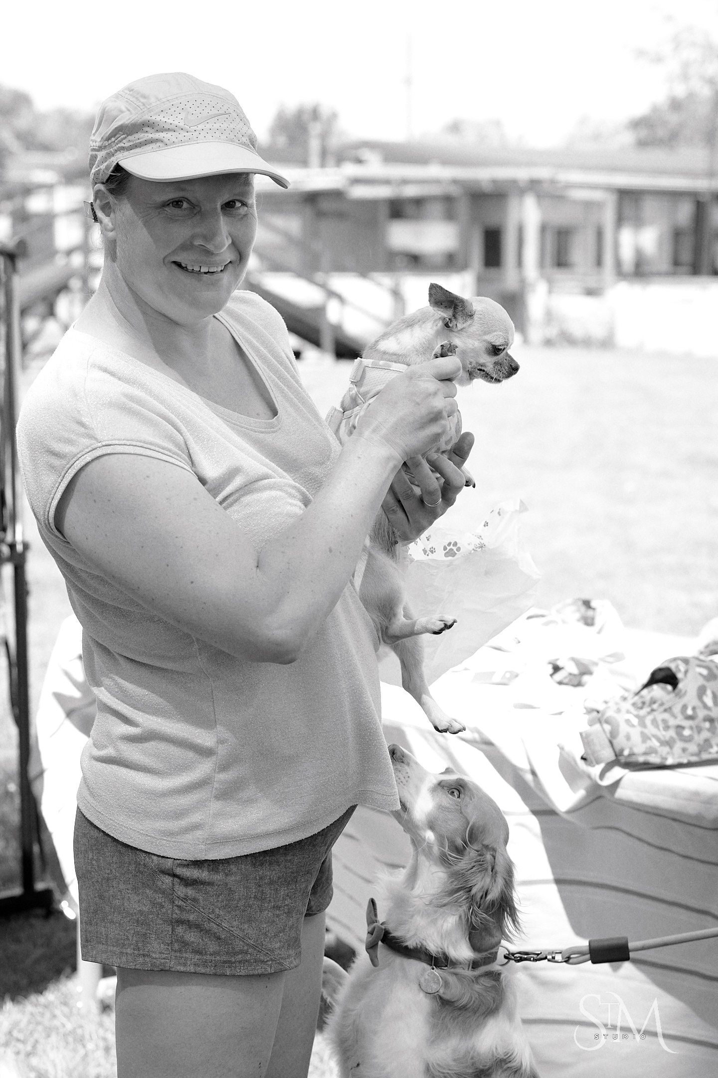 Woman in cap holds small Chihuahua while a Spaniel looks up curiously at an outdoor event.