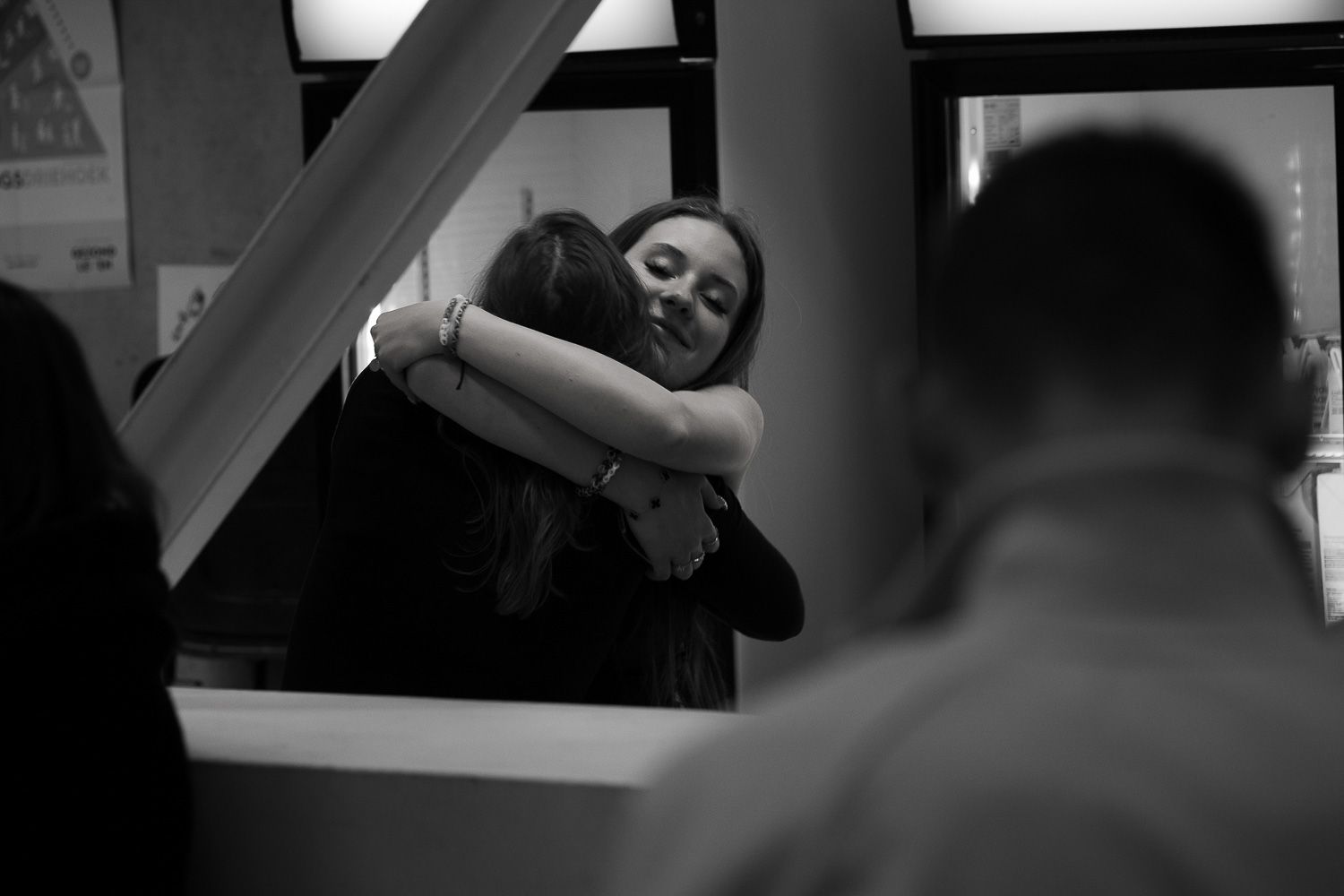 Two women share a warm embrace in a black and white indoor photo with blurred figures in the foreground.