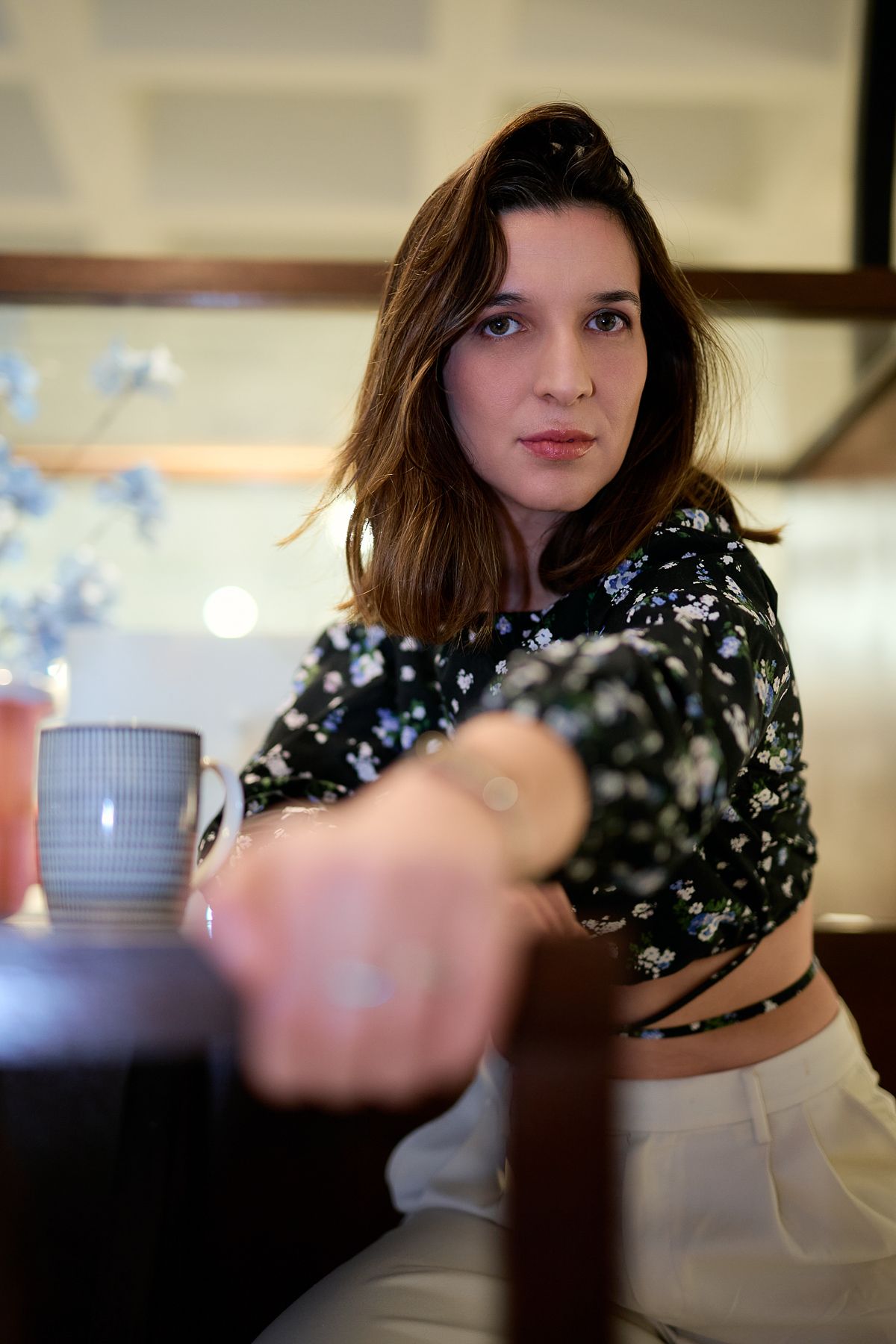 Young woman in floral crop top sitting at table with ceramic mug, gazing at camera in cozy indoor setting.