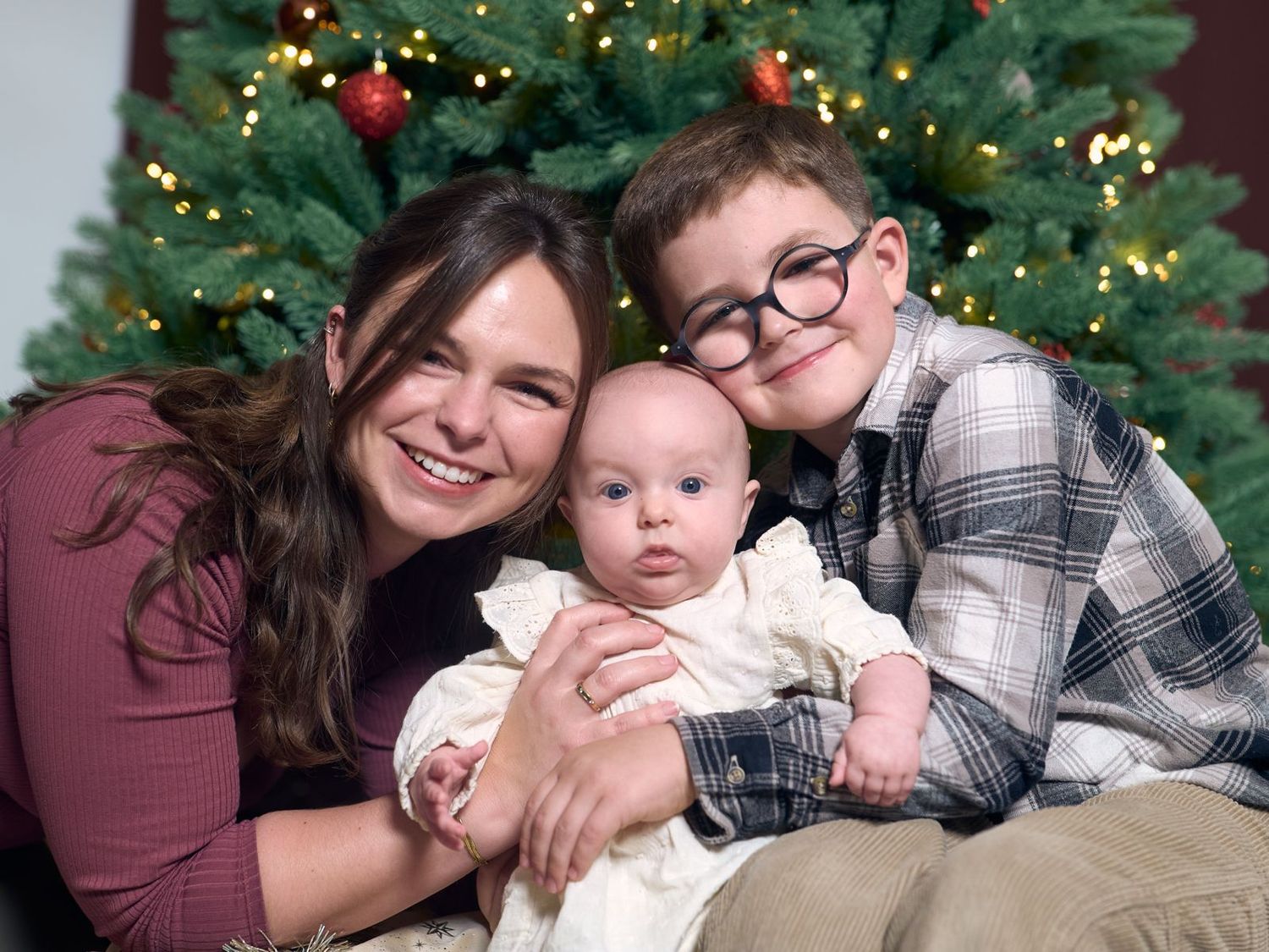Smiling mother with baby and young boy posing together in front of a festive Christmas tree.