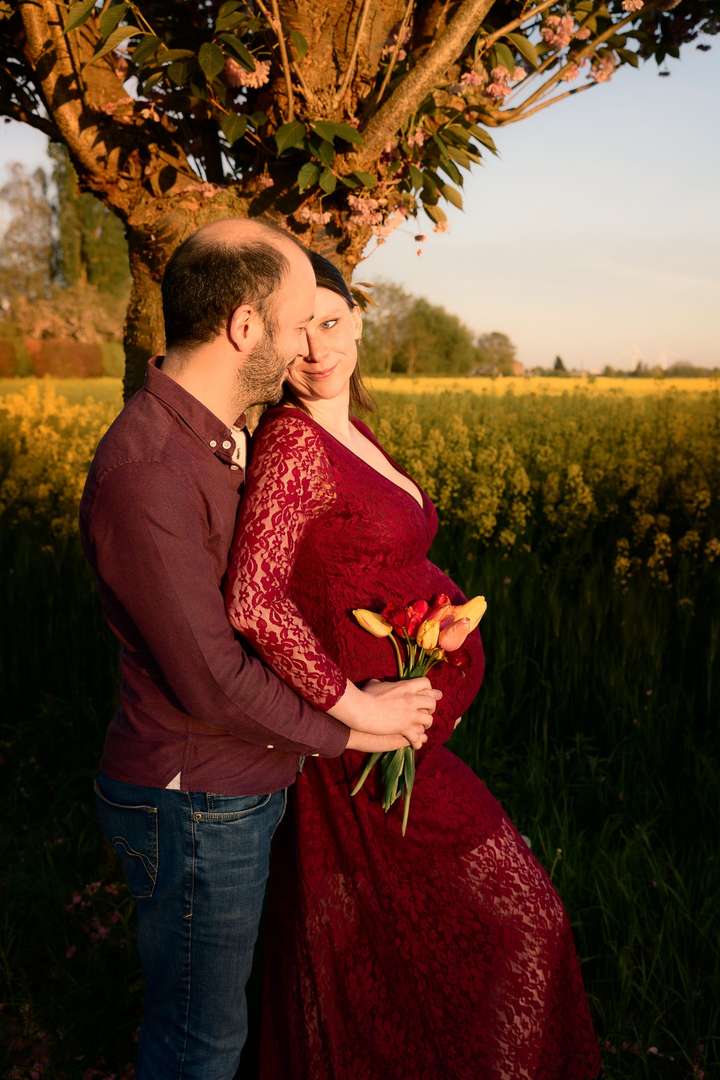 Couple embracing at golden hour near a tree, woman in red lace dress holding yellow and red tulips by a field.