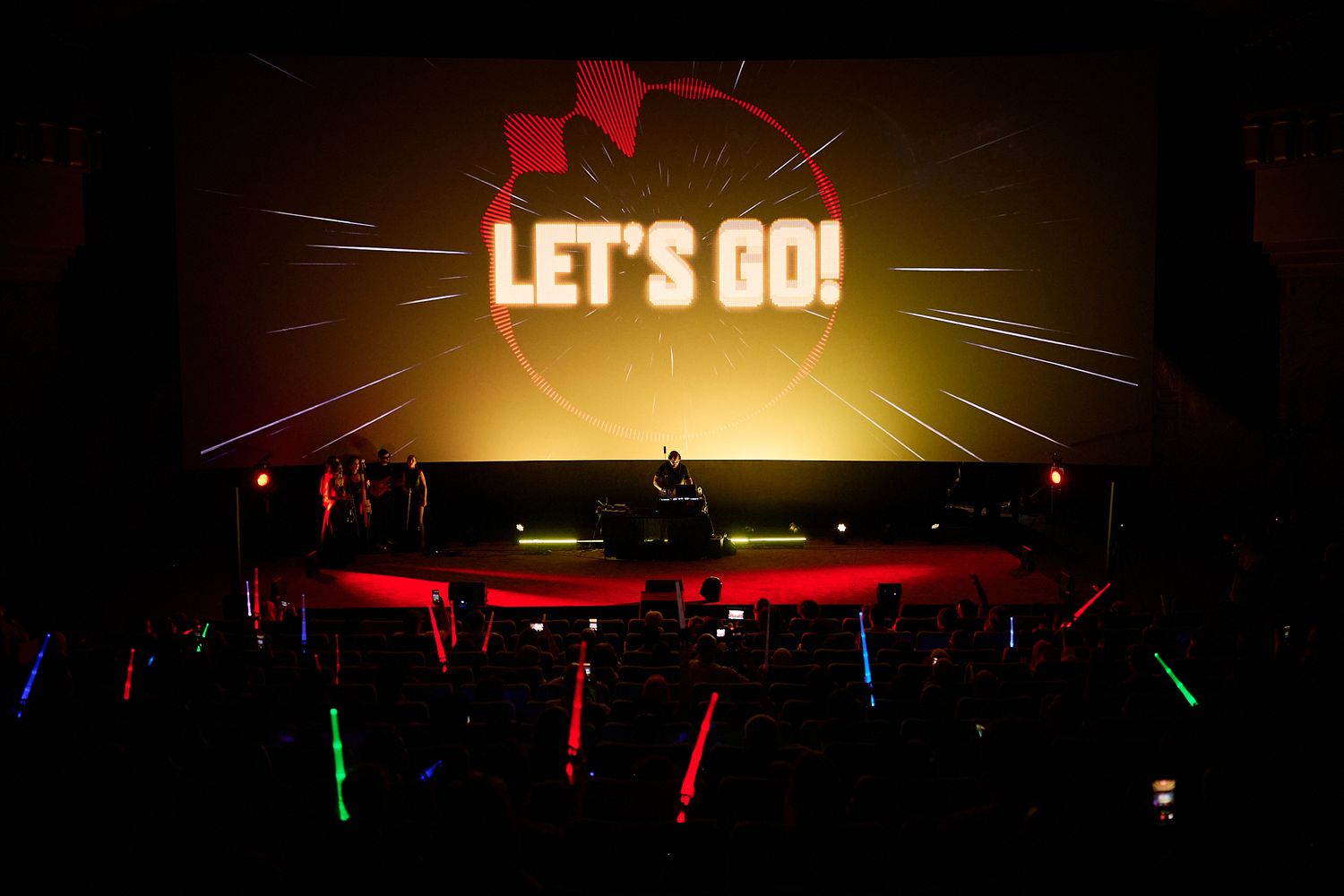 Concert stage with 'LET'S GO!' projected on screen, colorful glow sticks in crowd, red stage lighting.