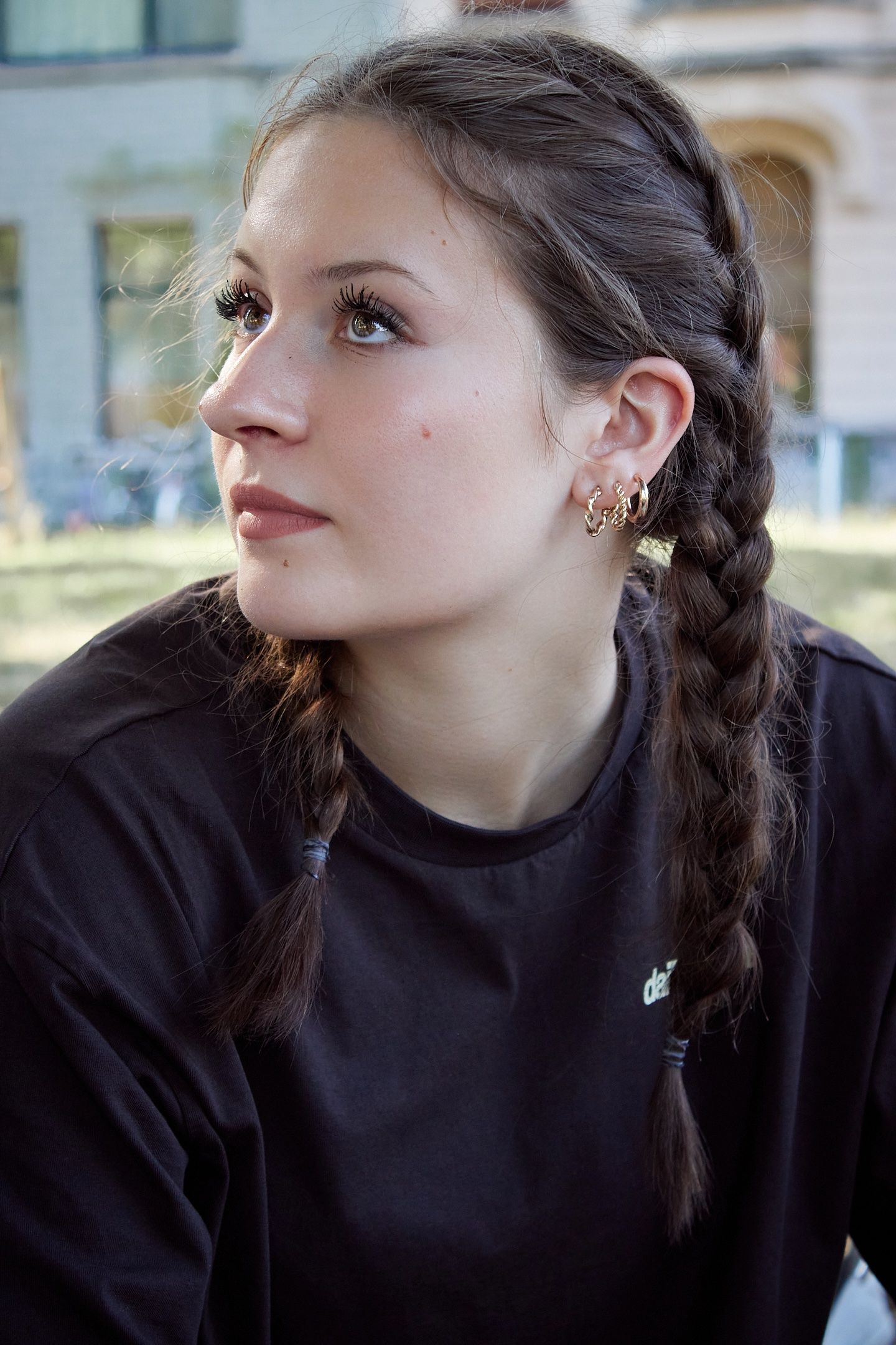 Young woman with dark braided hair and gold hoop earrings wearing a black shirt, gazing thoughtfully outdoors.