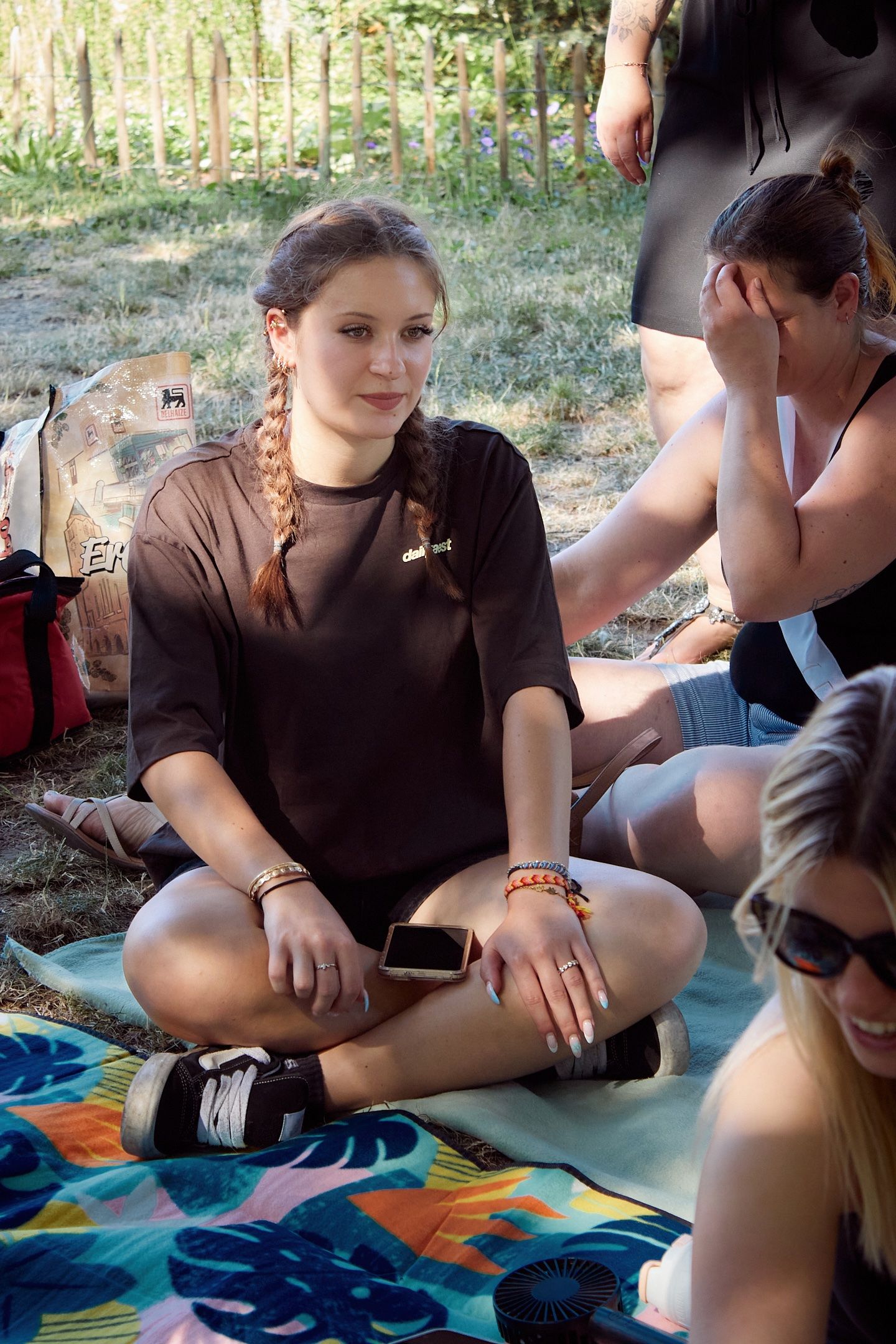 Young woman with braids sits cross-legged on colorful blanket outdoors, wearing brown t-shirt and bracelet.