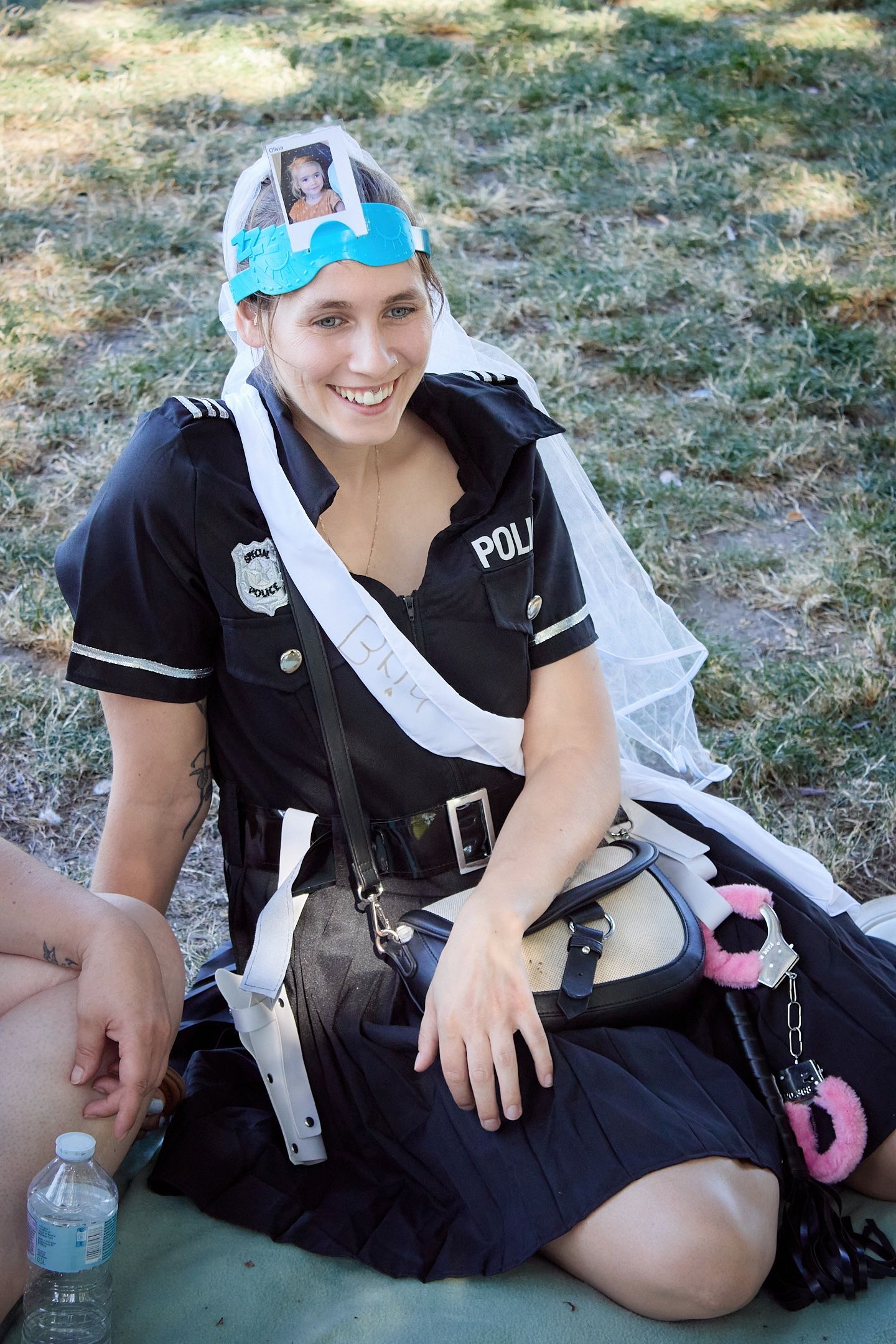 Woman in police costume with bridal veil and pink handcuffs smiling outdoors at a bachelorette party.