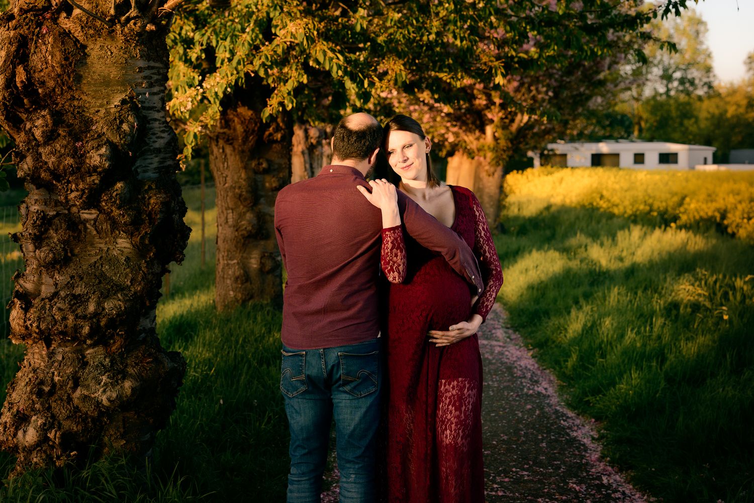 Pregnant couple embracing outdoors at golden hour, woman in burgundy maternity gown, surrounded by lush green trees.
