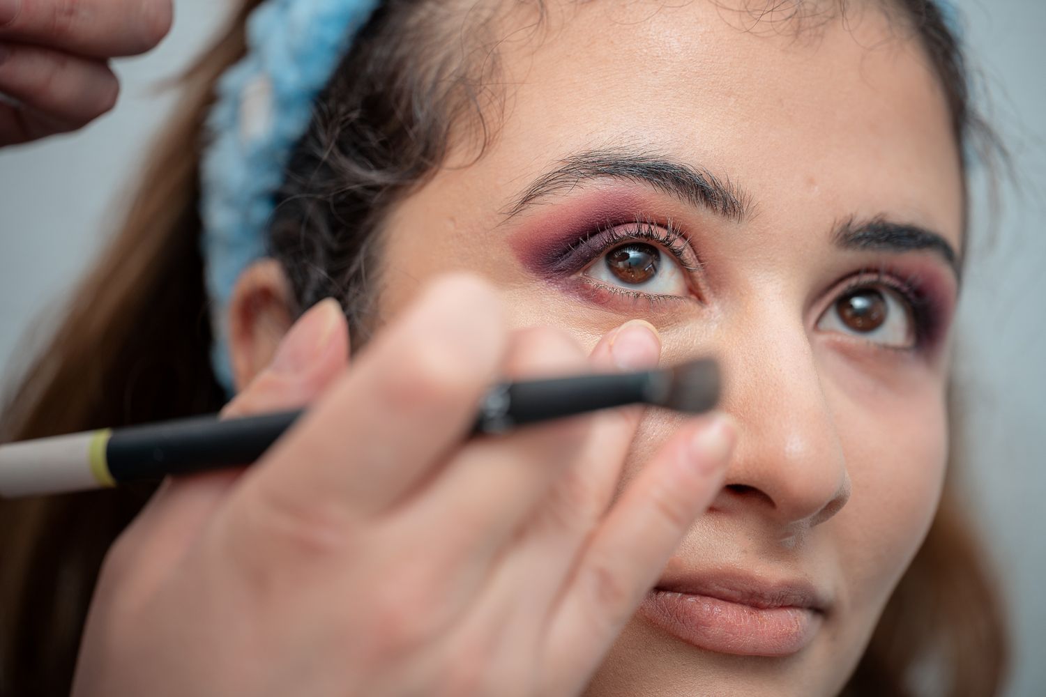 Makeup artist applying eyeliner to a young woman with pink eyeshadow and a blue headband.