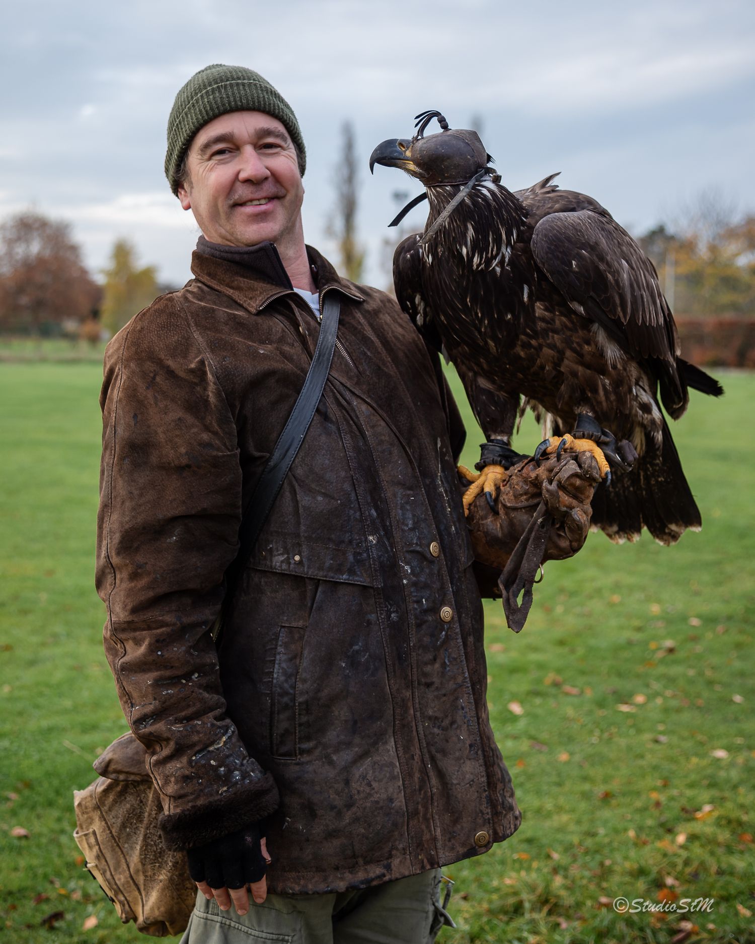 Falconer in brown leather jacket and green beanie holds a large eagle on gloved hand in autumn park.