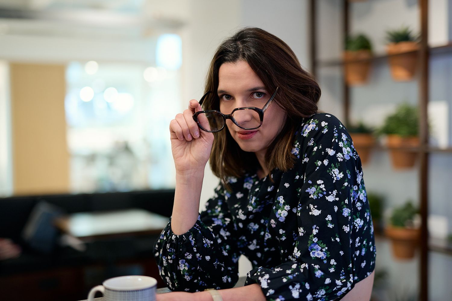 Woman in floral blouse adjusting glasses, smiling confidently at camera in modern office setting.