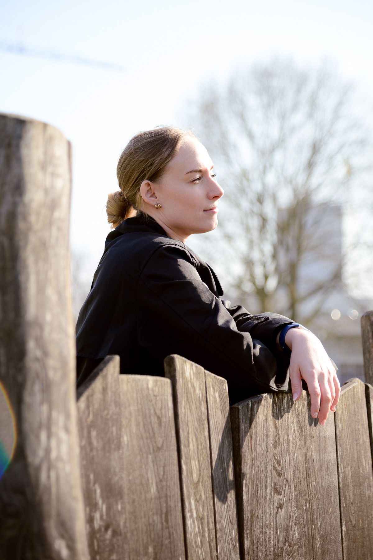 Young woman in black jacket leaning on rustic wooden fence outdoors, gazing thoughtfully into distance on sunny day.