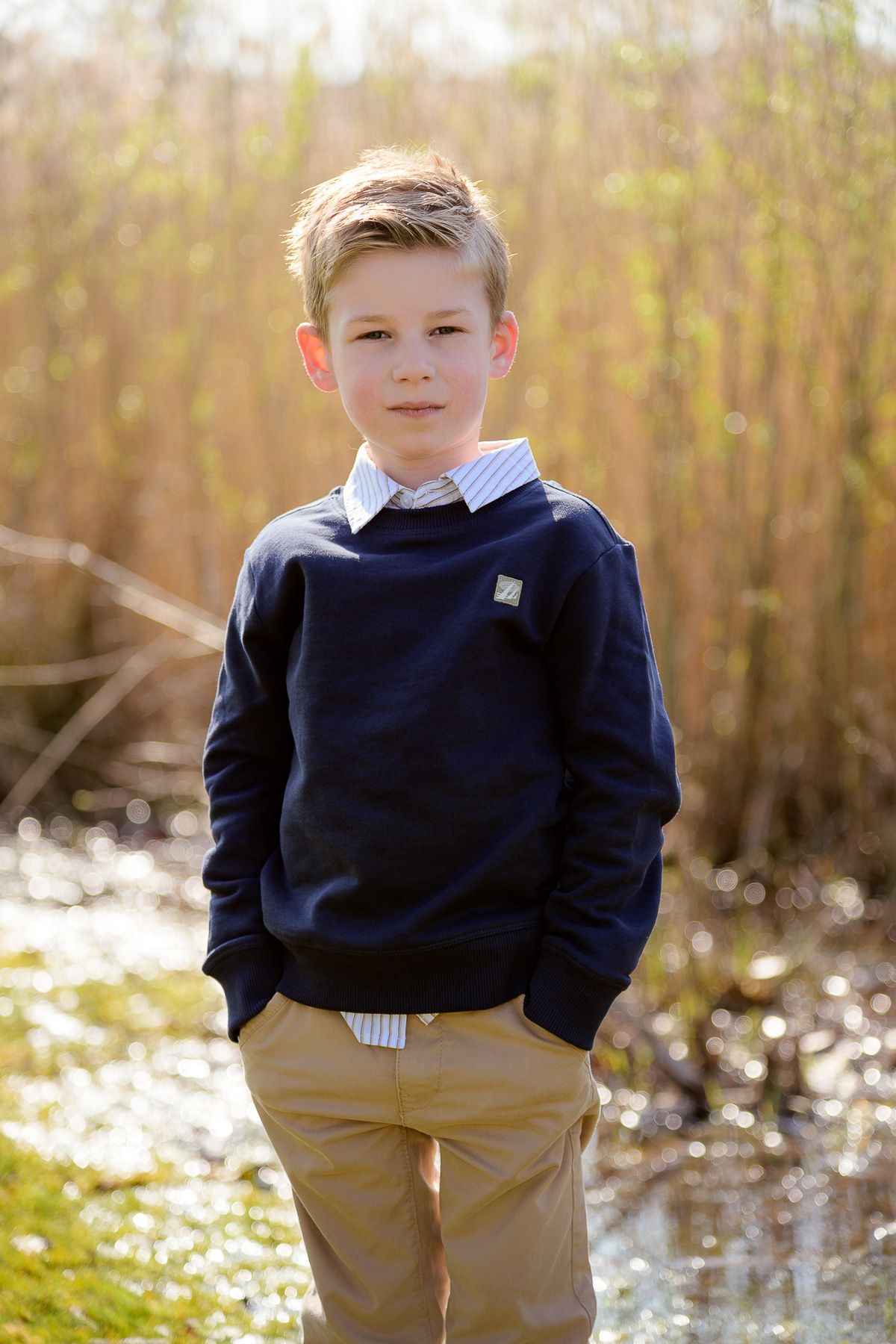 A young boy in a navy sweater and khaki pants stands outdoors near reeds and water on a sunny day.
