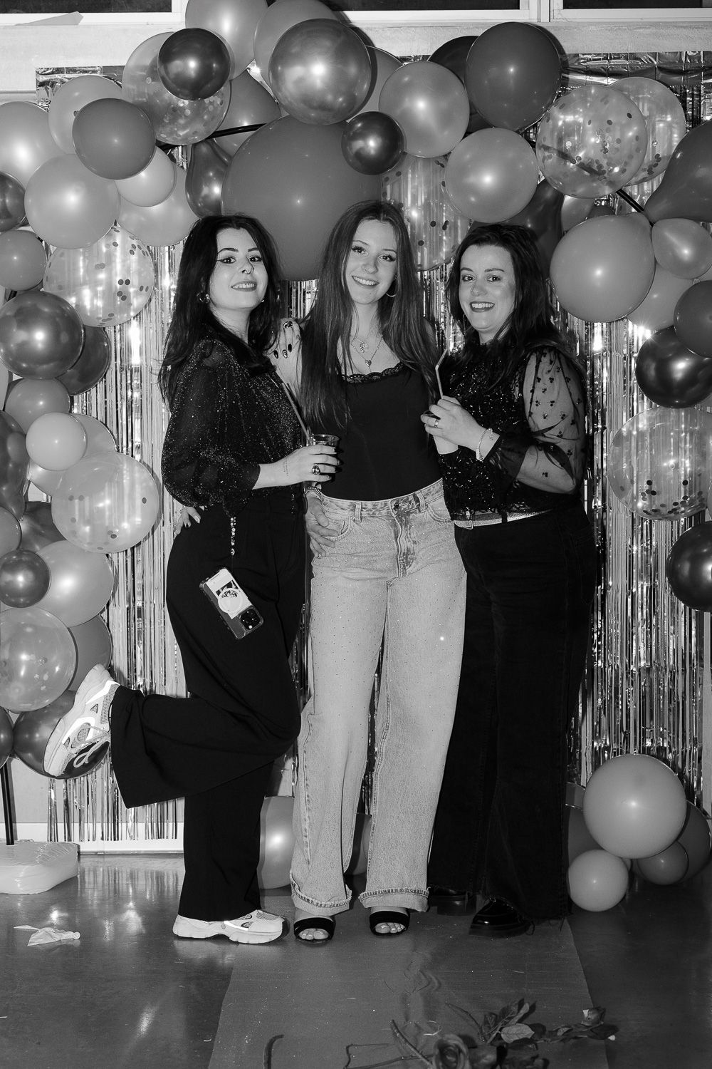 Three women posing together in front of a balloon arch at a party, dressed in elegant outfits.