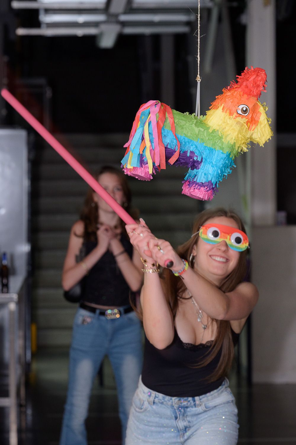 Girl wearing fun glasses swings a pink stick at a colorful donkey pinata at a party.