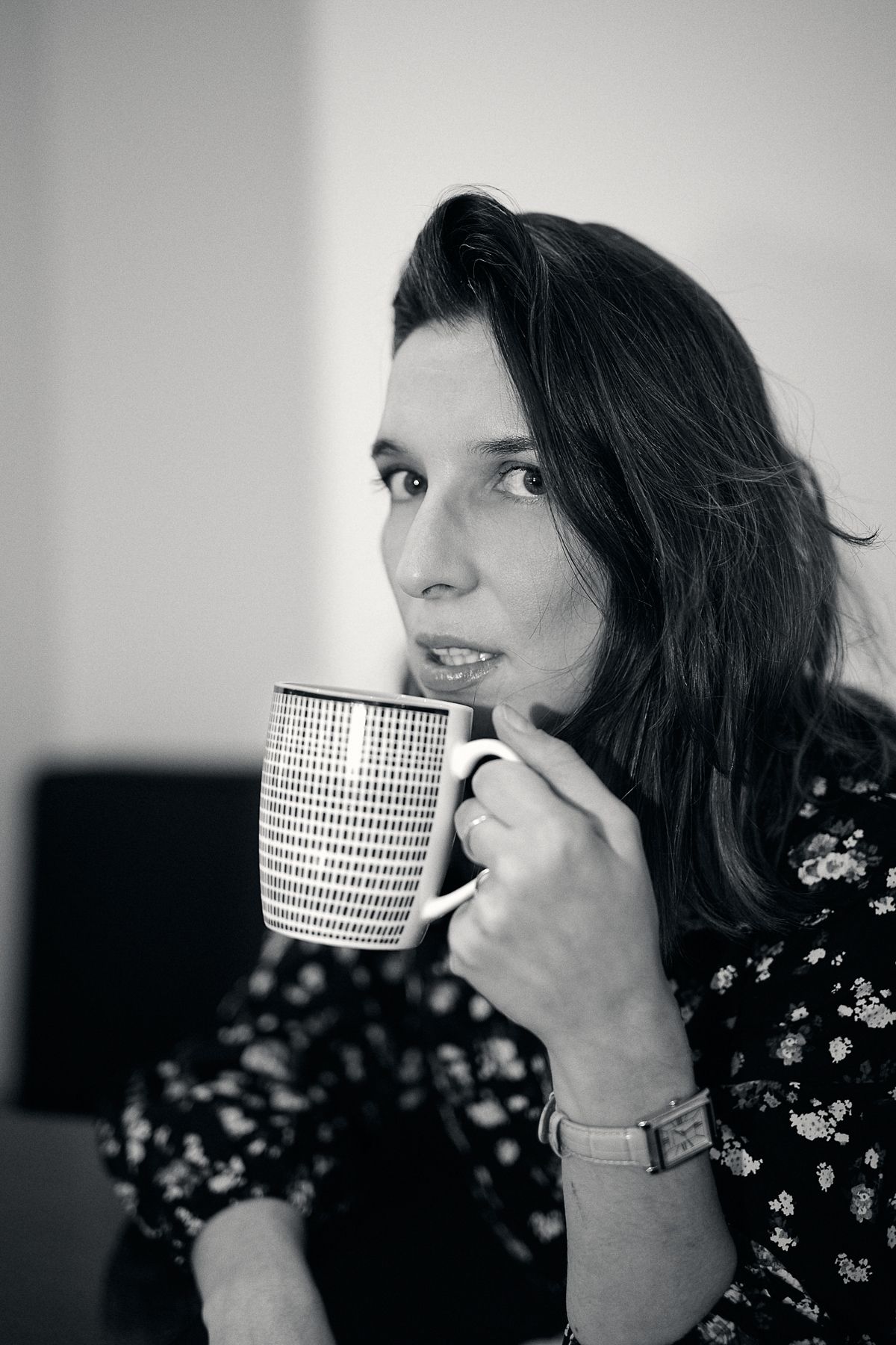 Black and white portrait of a woman holding a large patterned mug, wearing a floral dress and watch.