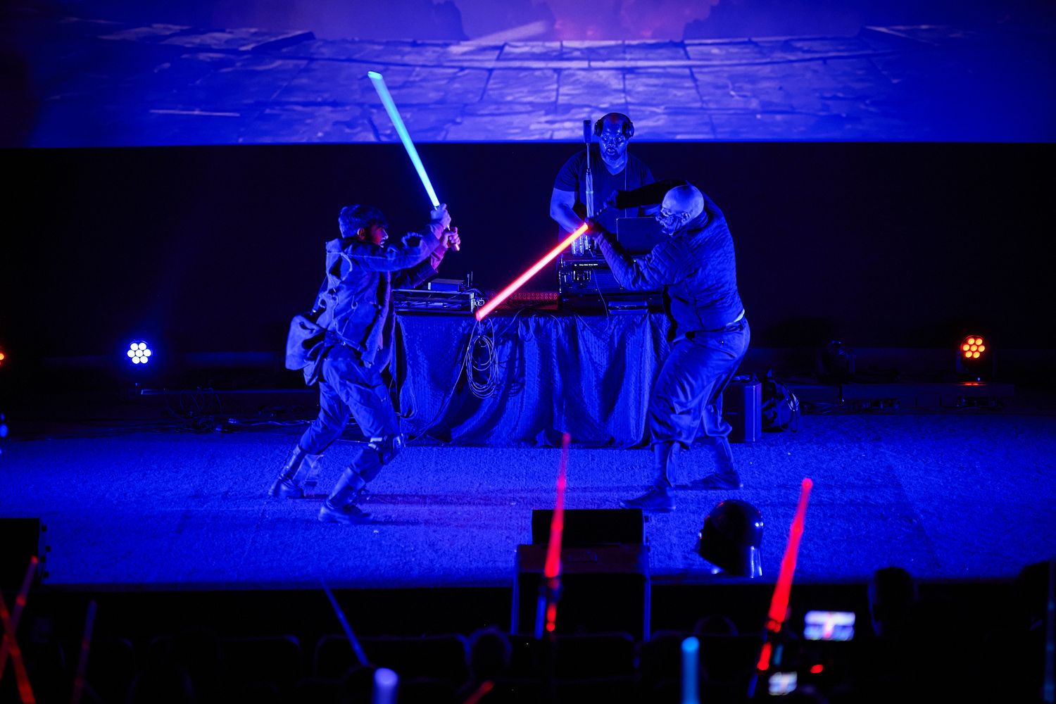 Two performers duel with glowing lightsabers on a dark stage bathed in blue light at a Star Wars themed event.