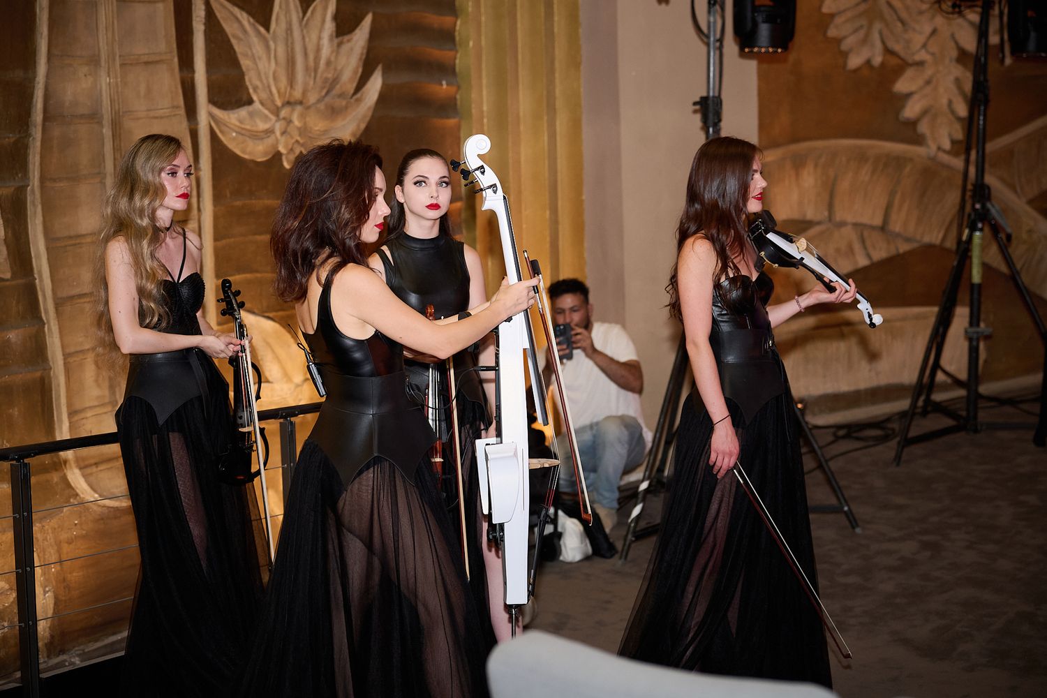 Four women in elegant black gowns perform with string instruments at a formal concert event.