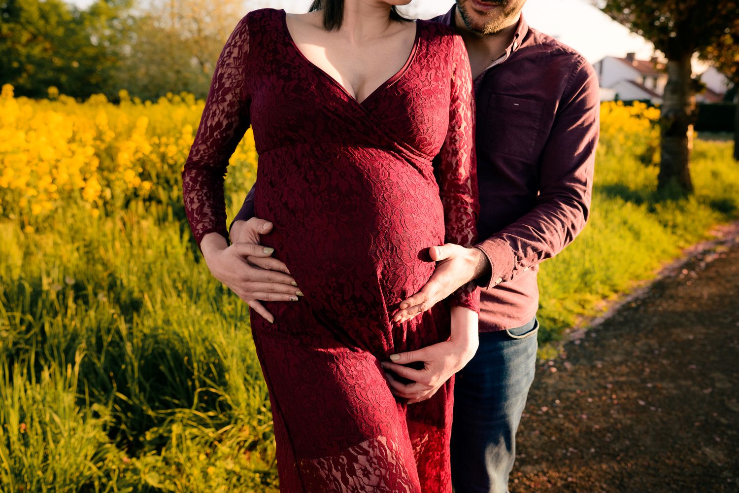 Pregnant woman in red lace dress with partner embracing her belly outdoors in golden sunlight near yellow flowers.