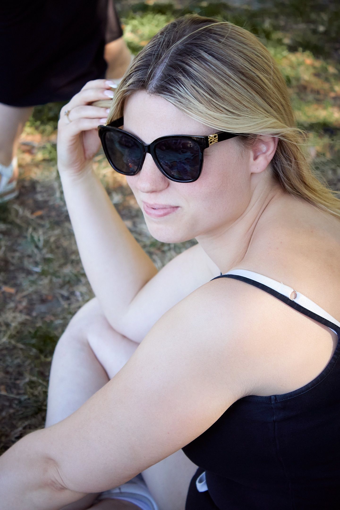 Young blonde woman wearing black sunglasses and tank top sitting outdoors, posing with hand raised to her head.