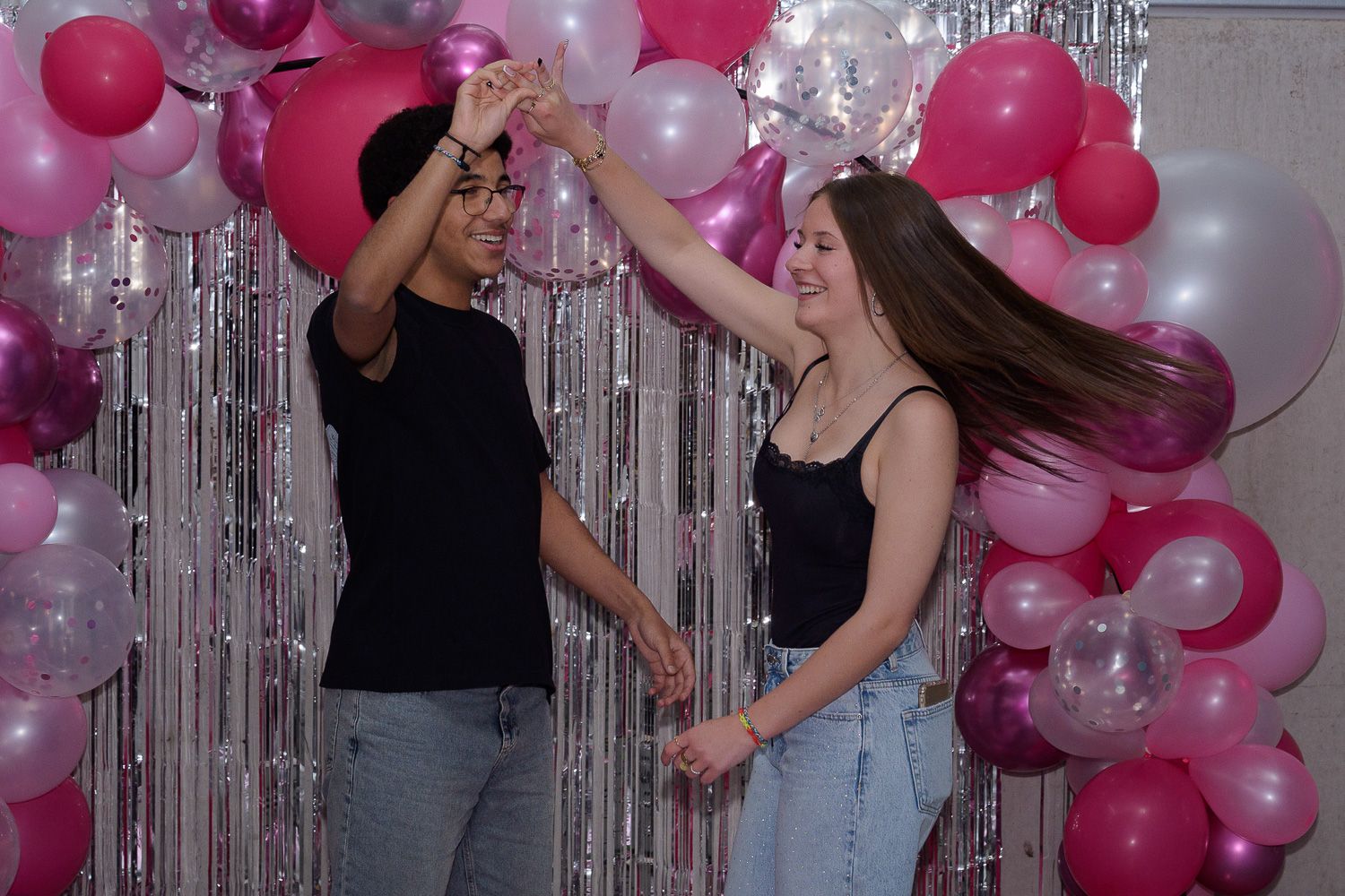Two teens dancing joyfully in front of pink and white balloon garland with silver tinsel curtain backdrop.