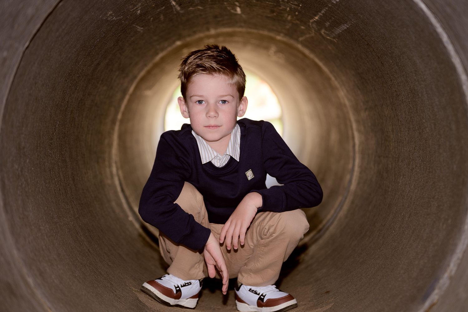 Young boy crouching inside a large concrete tunnel, wearing navy blazer and khaki pants.
