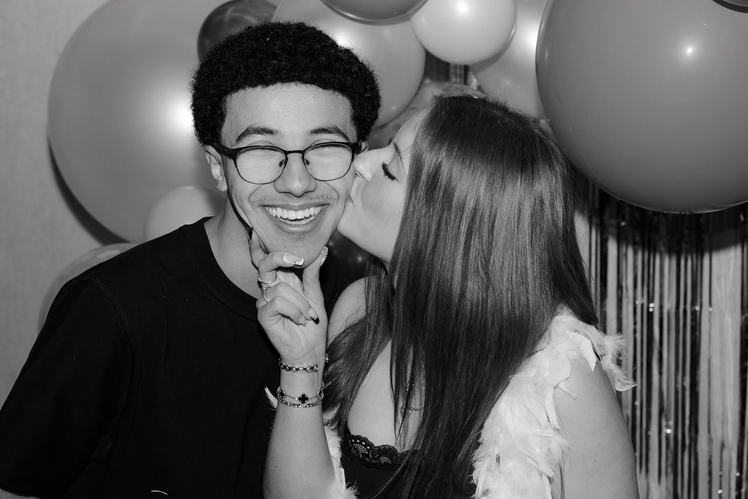 Young couple celebrating at a party, woman kissing man on cheek with balloons in black and white photo.