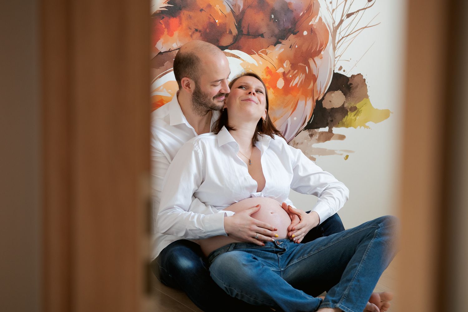 Couple sharing a tender moment, pregnant woman cradled by partner against a colorful floral wall mural.
