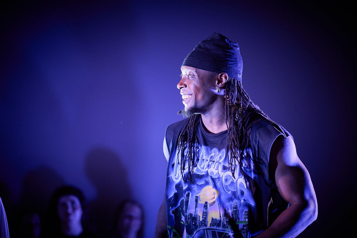 A muscular performer with dreadlocks and headband rocks a sleeveless band tee on a purple-lit stage.