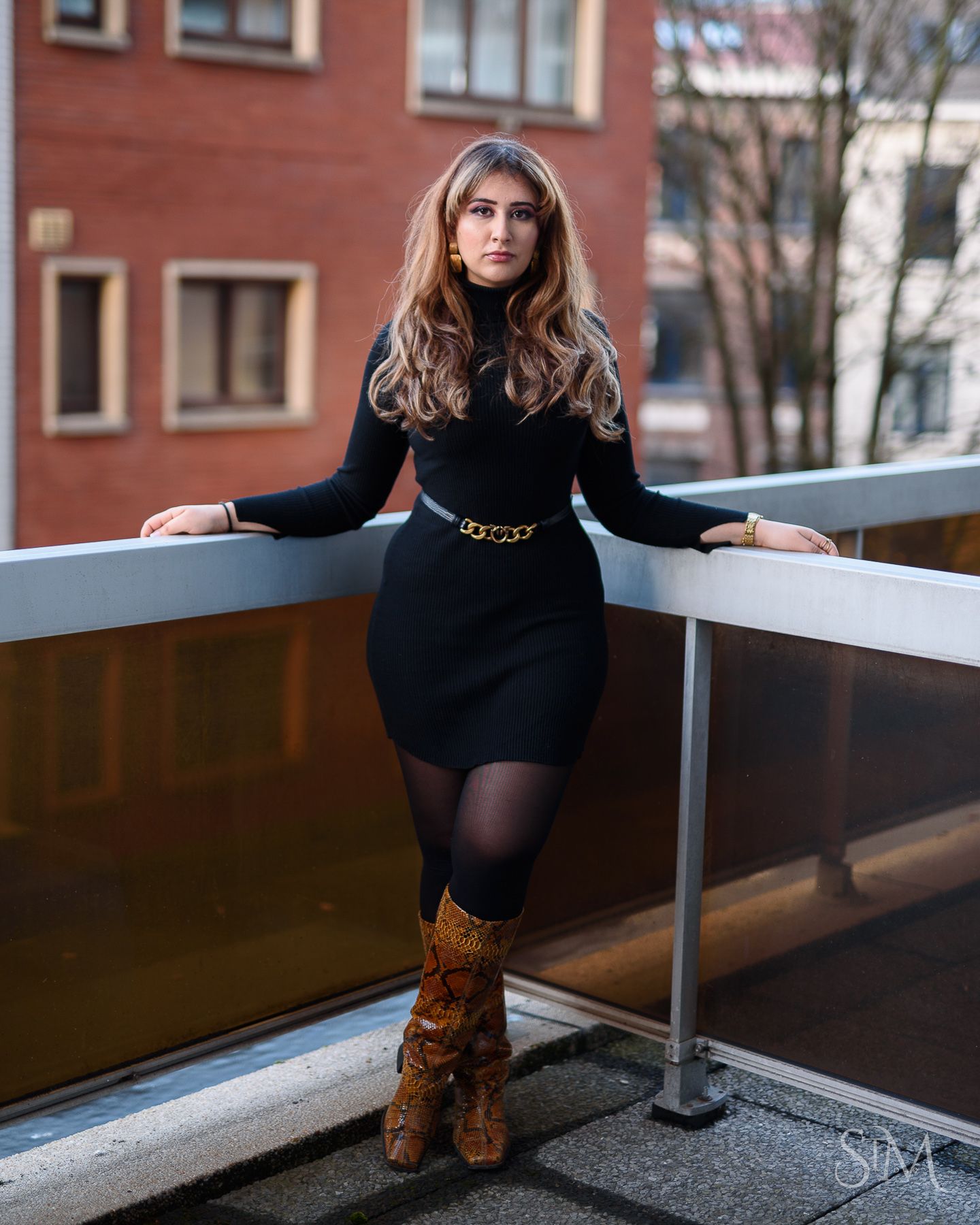 Woman in black turtleneck dress, gold chain belt, sheer tights, and brown snake-print boots posing on urban balcony.