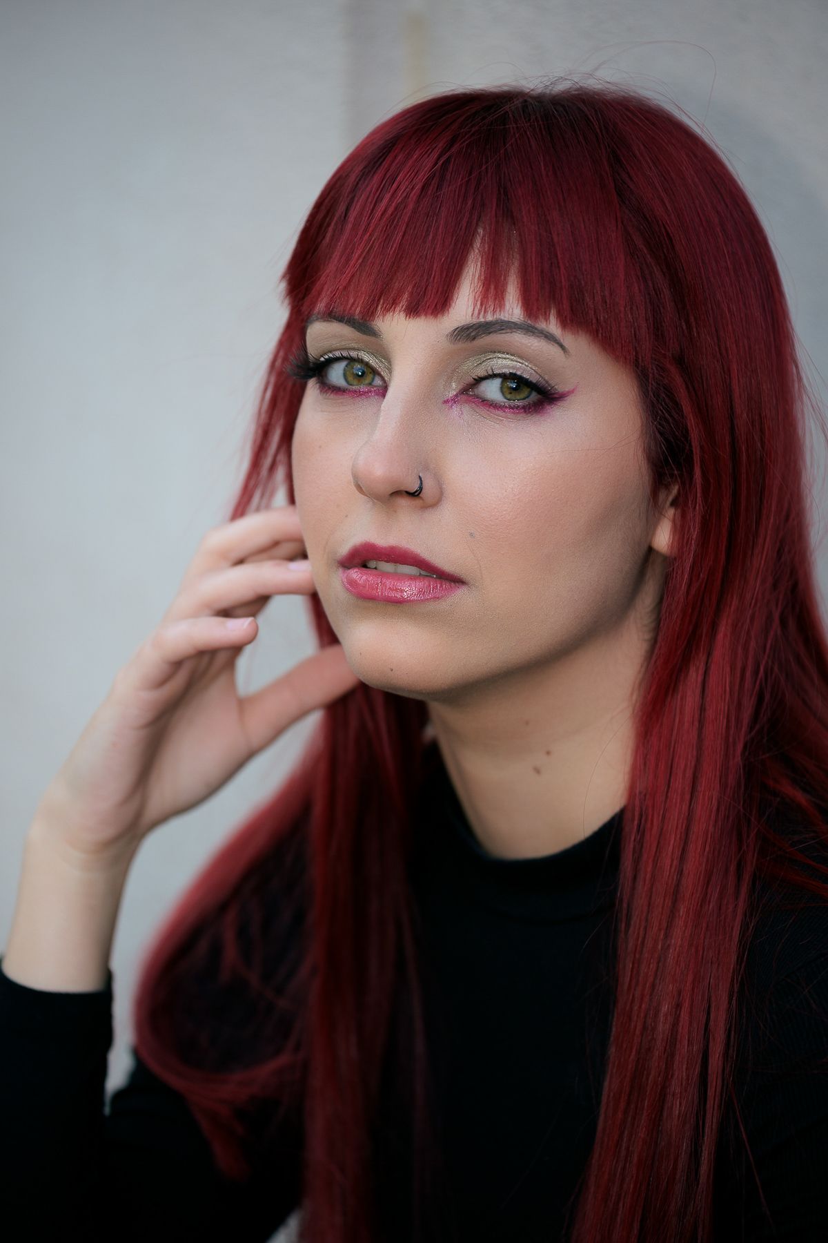 Portrait of a woman with vibrant red hair and colorful eye makeup against a white wall.