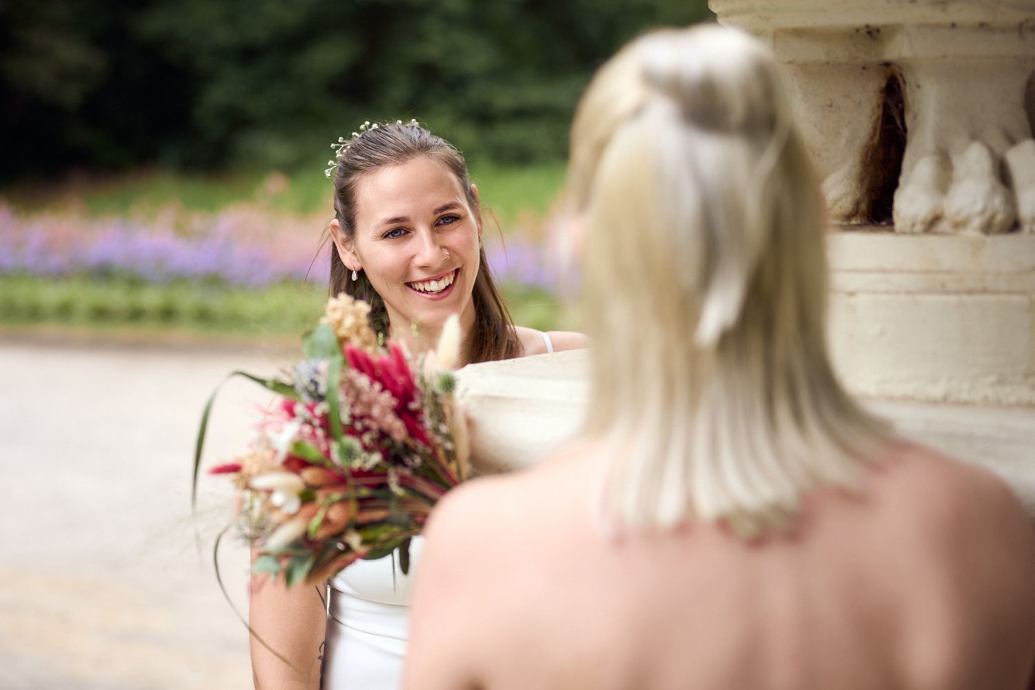 Two brides in white dresses share a joyful moment outdoors, one holding a colorful bouquet near a stone fountain.