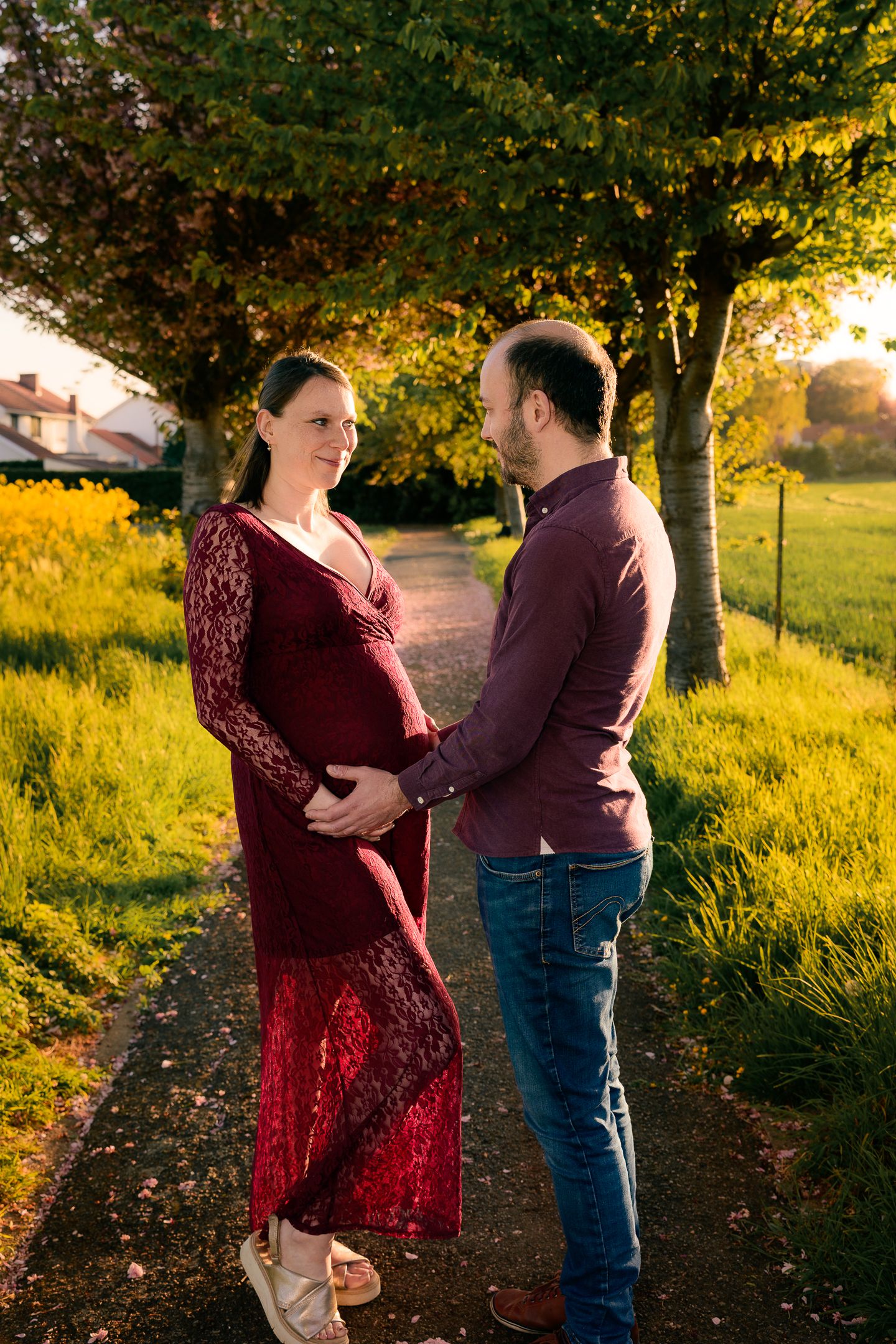 Pregnant couple holding baby bump outdoors at golden hour, woman in red lace dress, man in burgundy shirt and jeans.