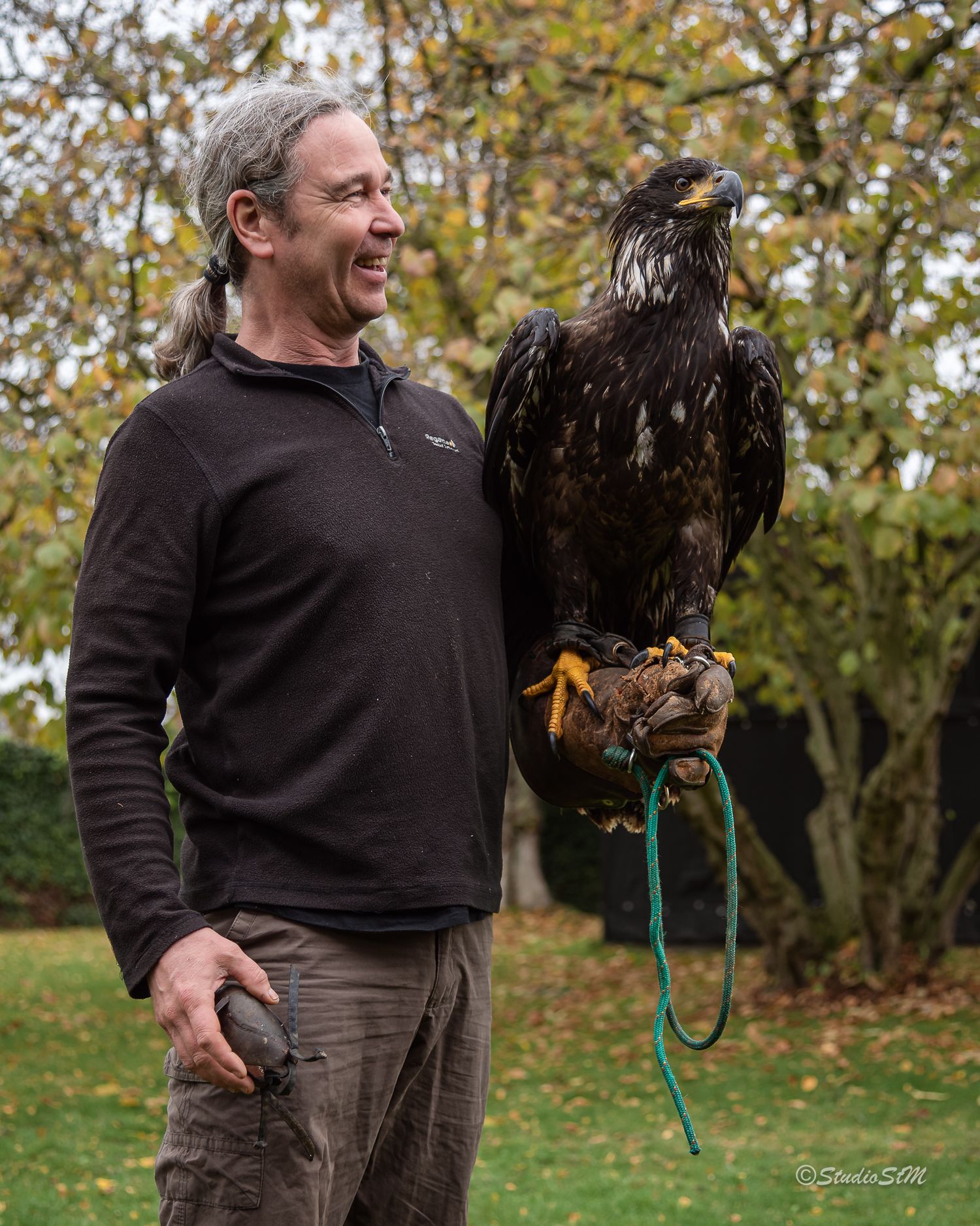 Man smiling while holding a large eagle on his gloved hand outdoors in a garden with autumn trees.