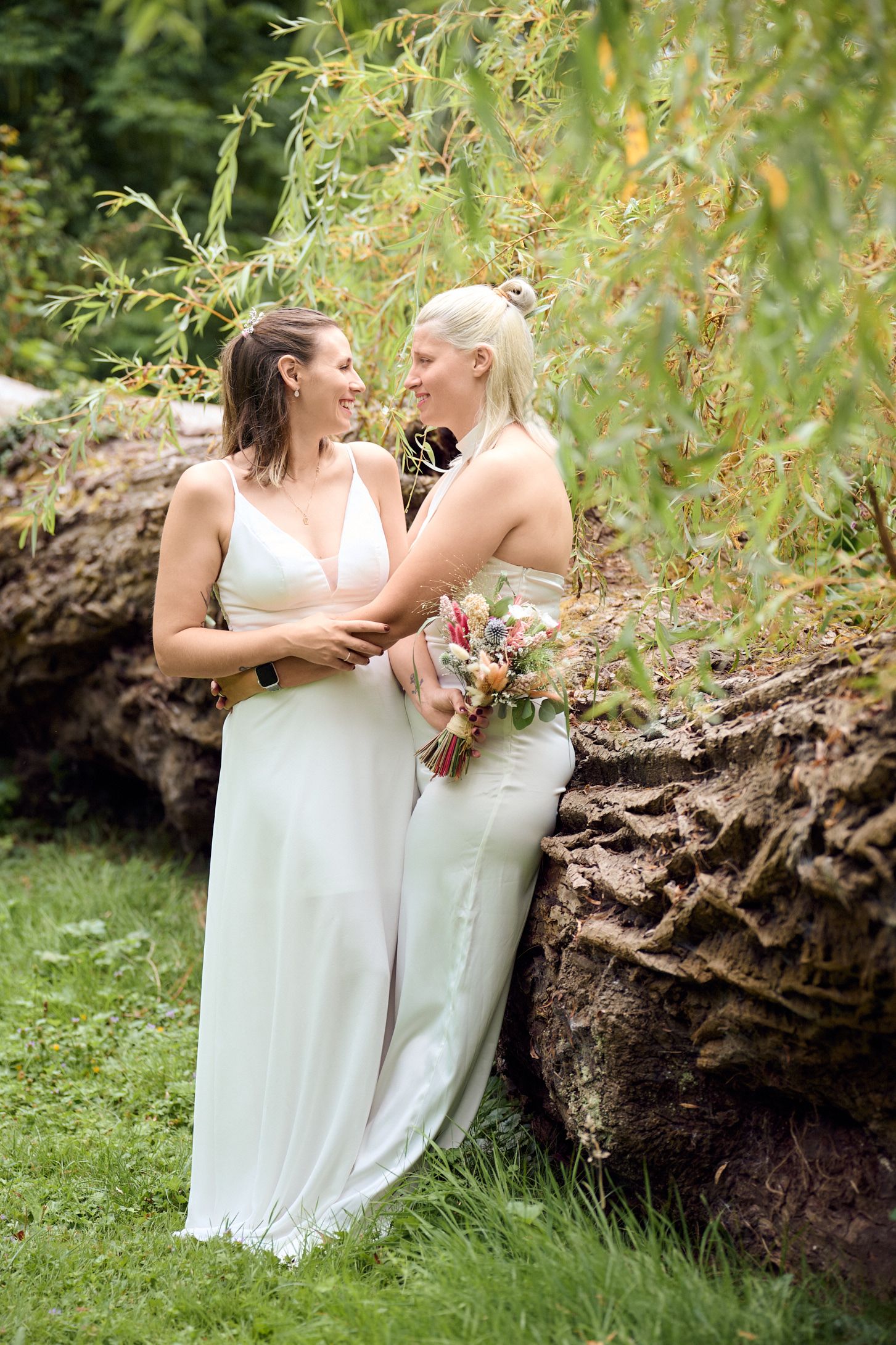 Two brides in white gowns share a joyful moment outdoors, one holding a wildflower bouquet near a mossy log.
