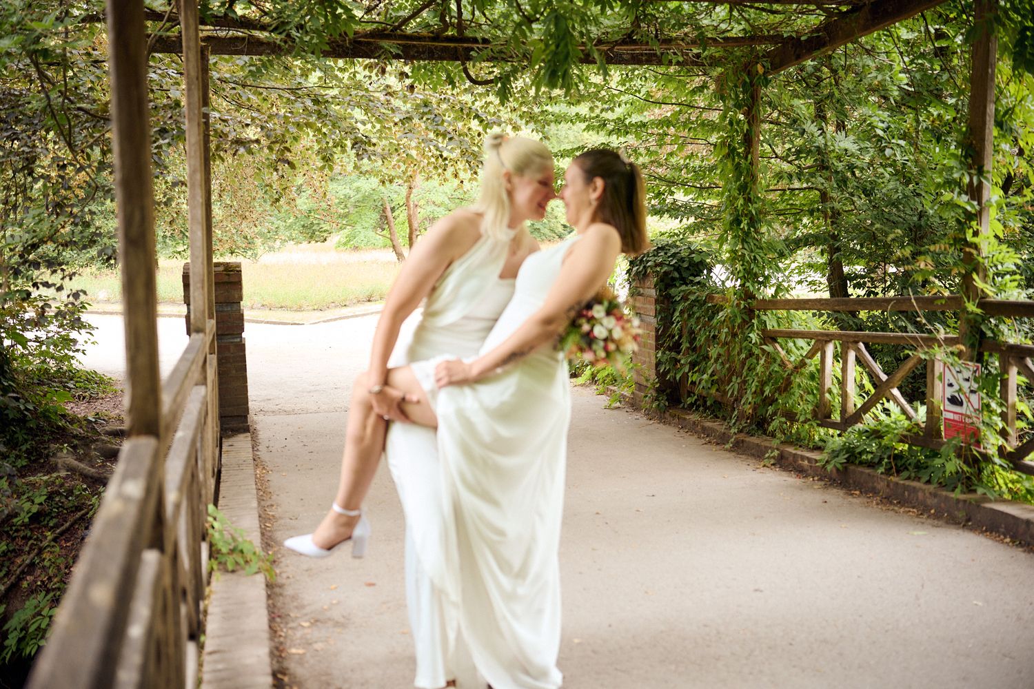 Two brides in white dresses share a romantic moment on a garden bridge surrounded by lush greenery.