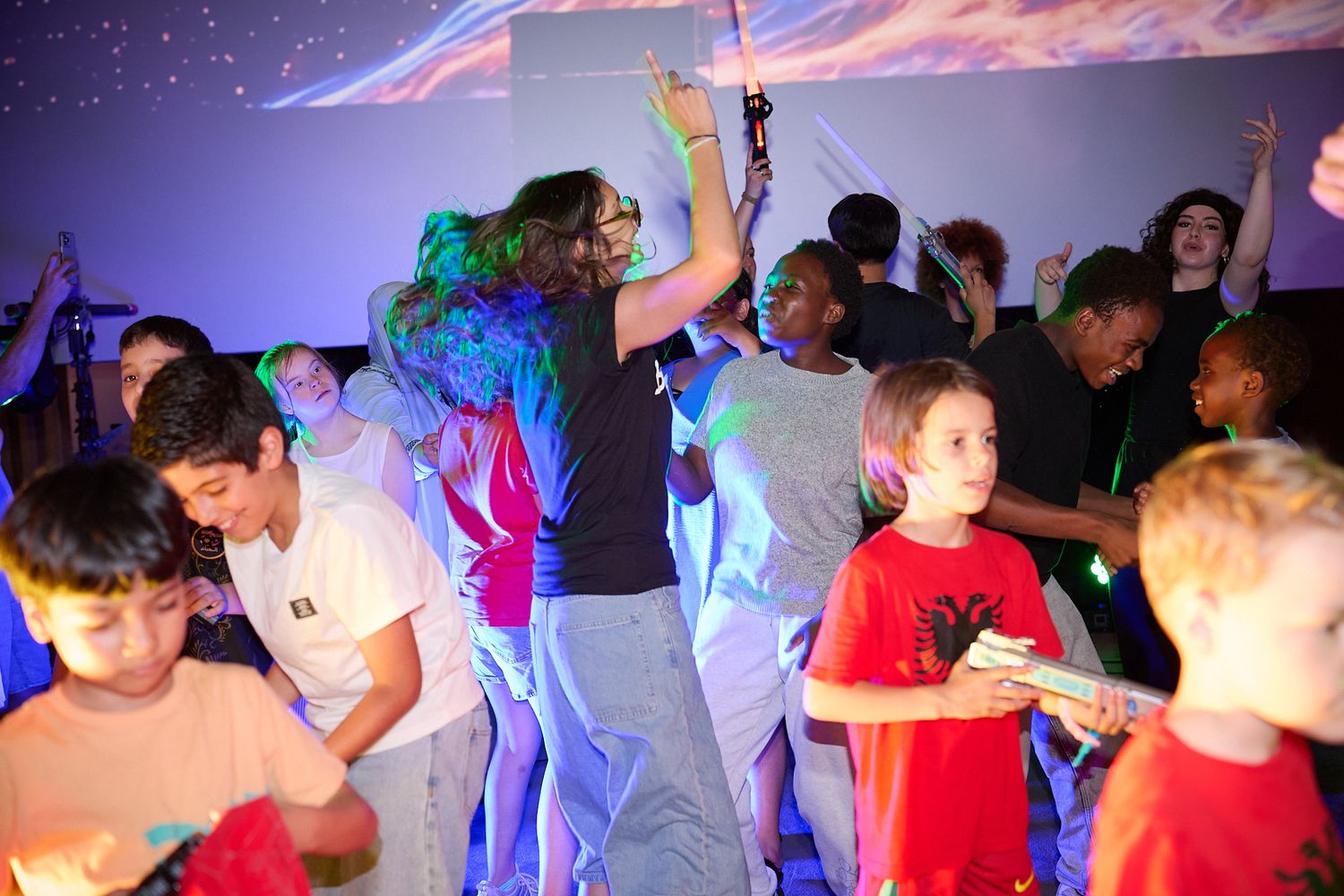 Children and adults dancing together at a colorful indoor party with vibrant lighting and music.