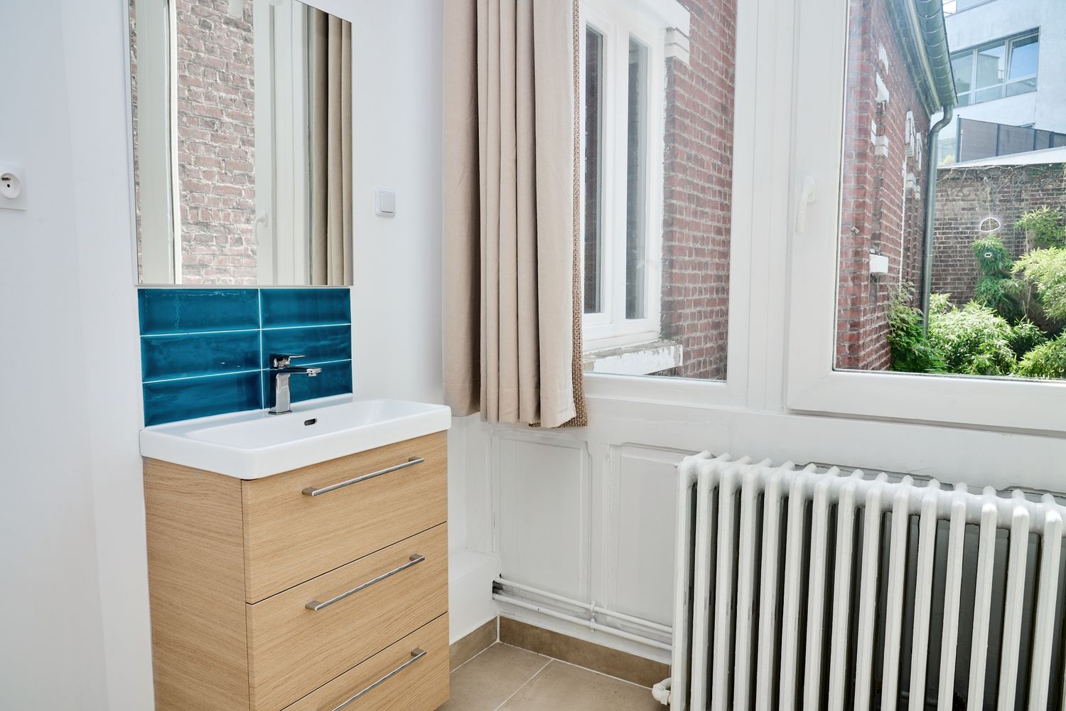 Modern bathroom vanity with wooden cabinet, white sink, blue tile backsplash, mirror, and white radiator near a bright window.