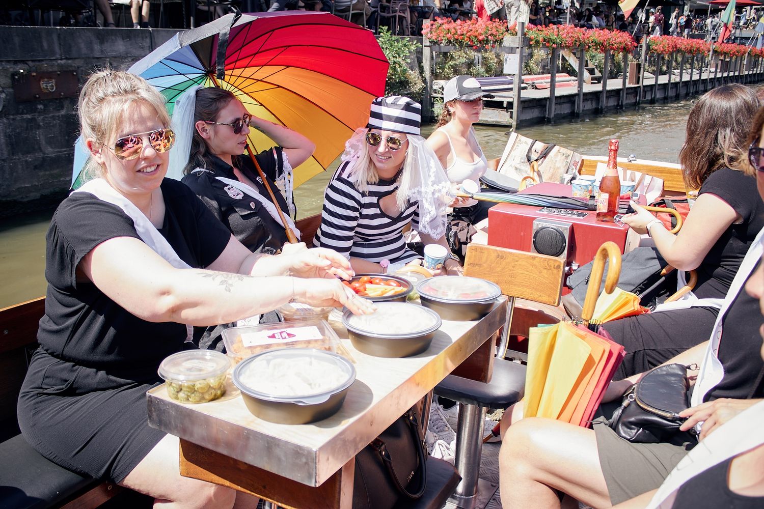 Group of women dining on a boat at a sunny floating market, enjoying food under colorful umbrellas.