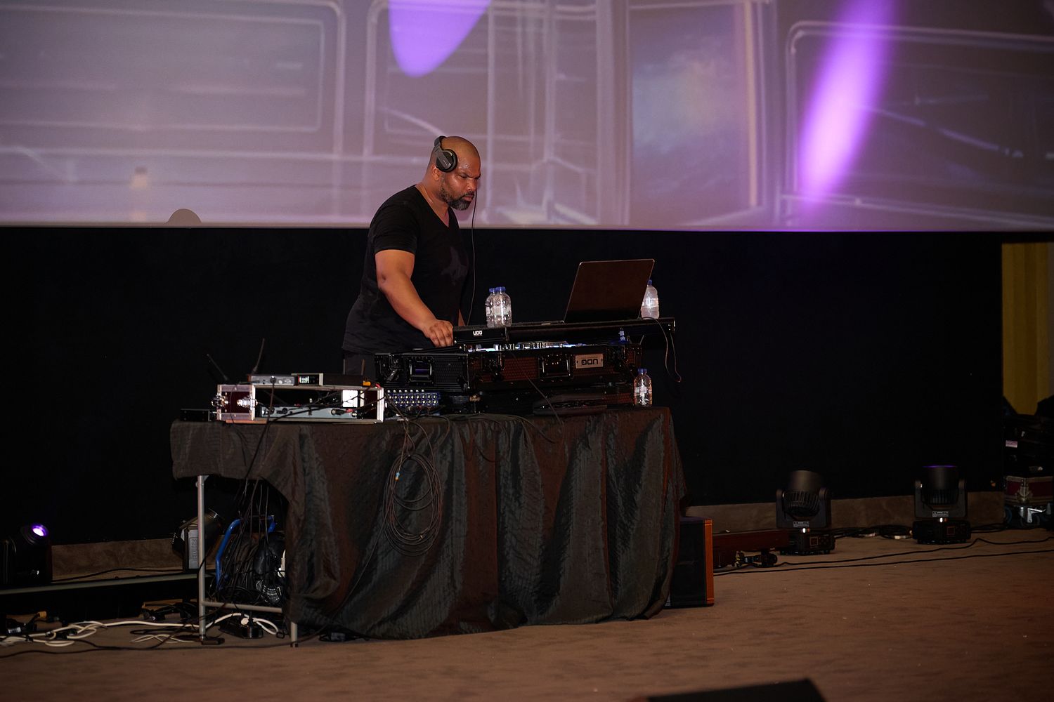 DJ performing on stage at a live event, mixing music on turntables with a large projection screen in the background.