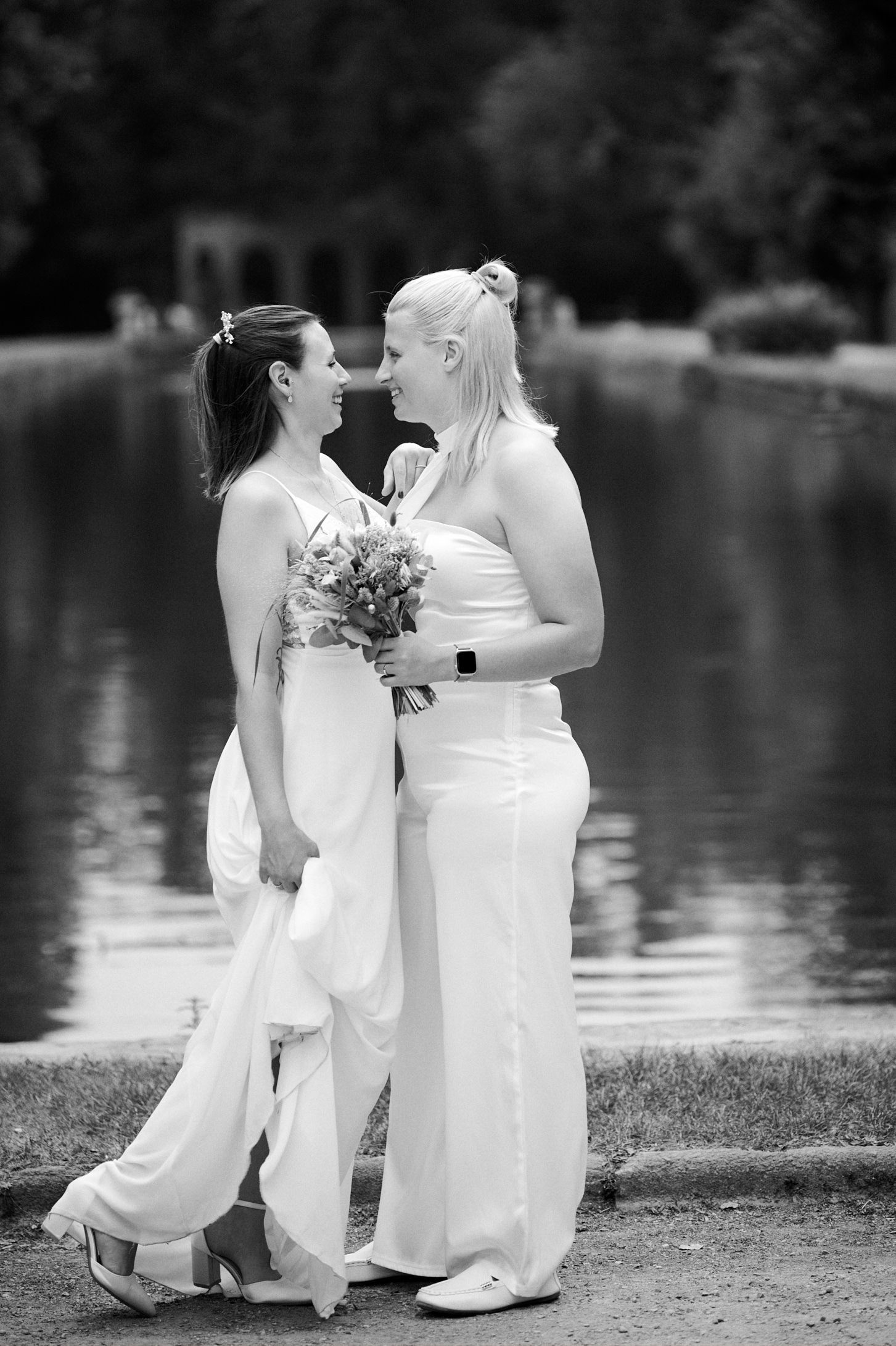 Two brides in white dresses share romantic moment by lake in black and white wedding photo.