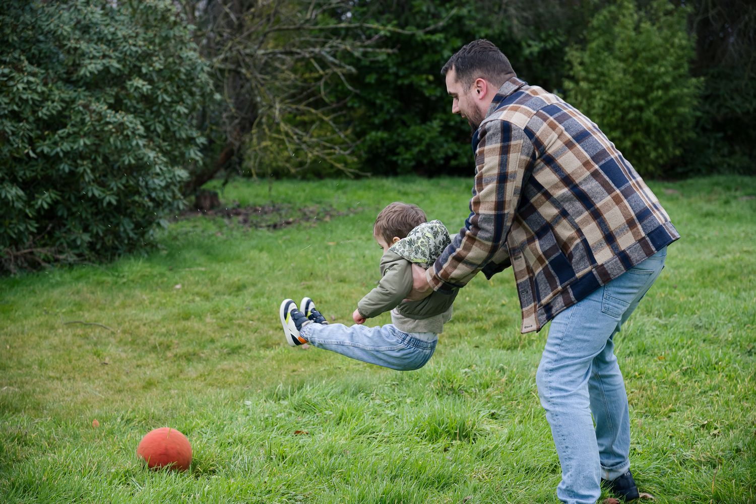 Father in plaid shirt playing football with his young child on a green lawn outdoors.