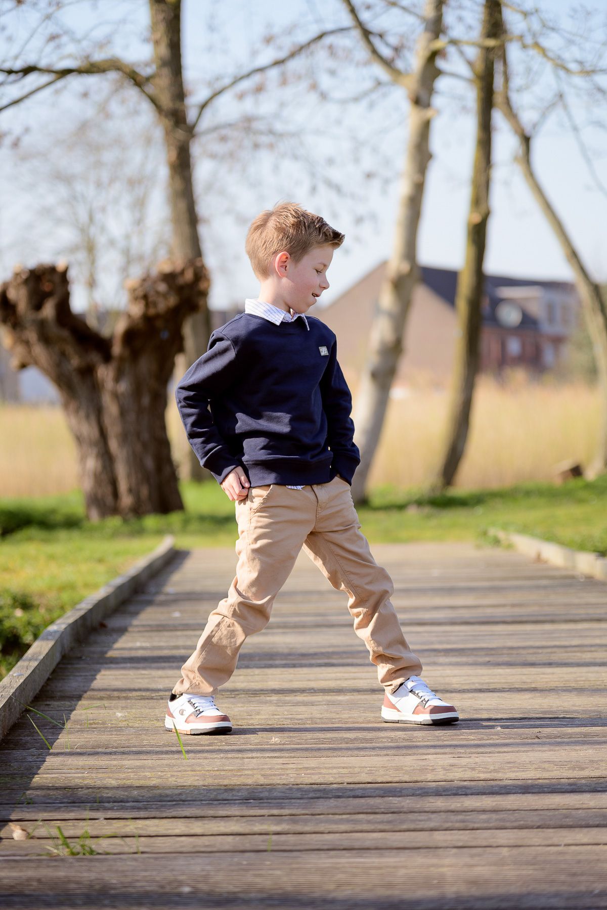 Boy in navy sweater and khaki pants walking on wooden boardwalk path in sunny park with bare trees.