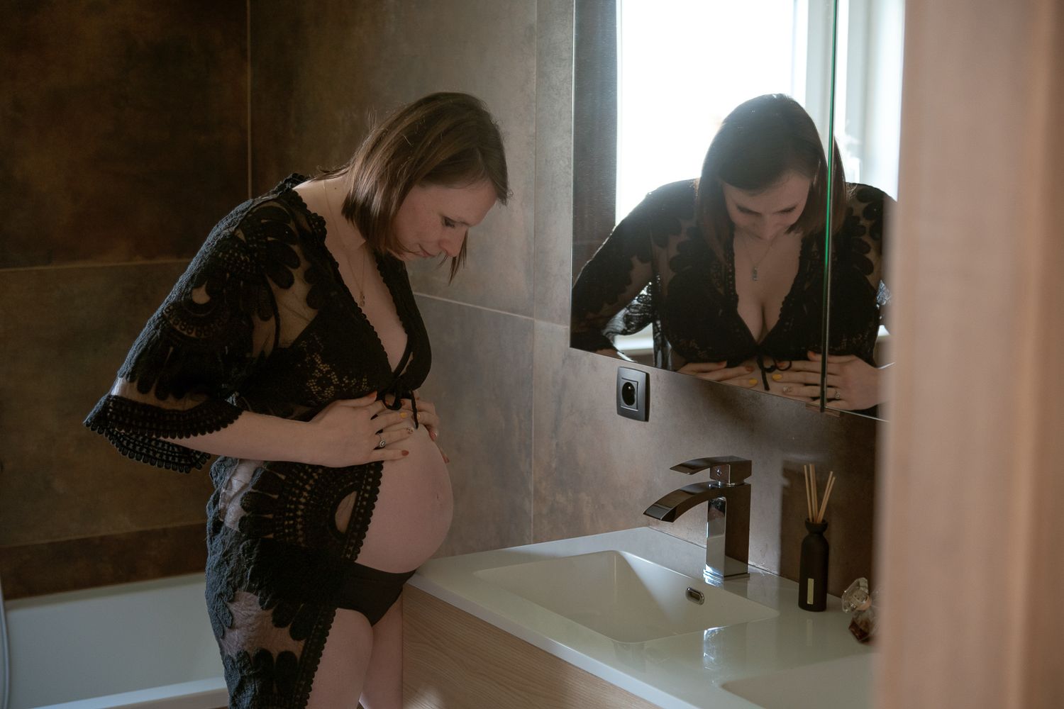 Pregnant woman in black lace robe examining baby bump in bathroom mirror, maternity boudoir photography.