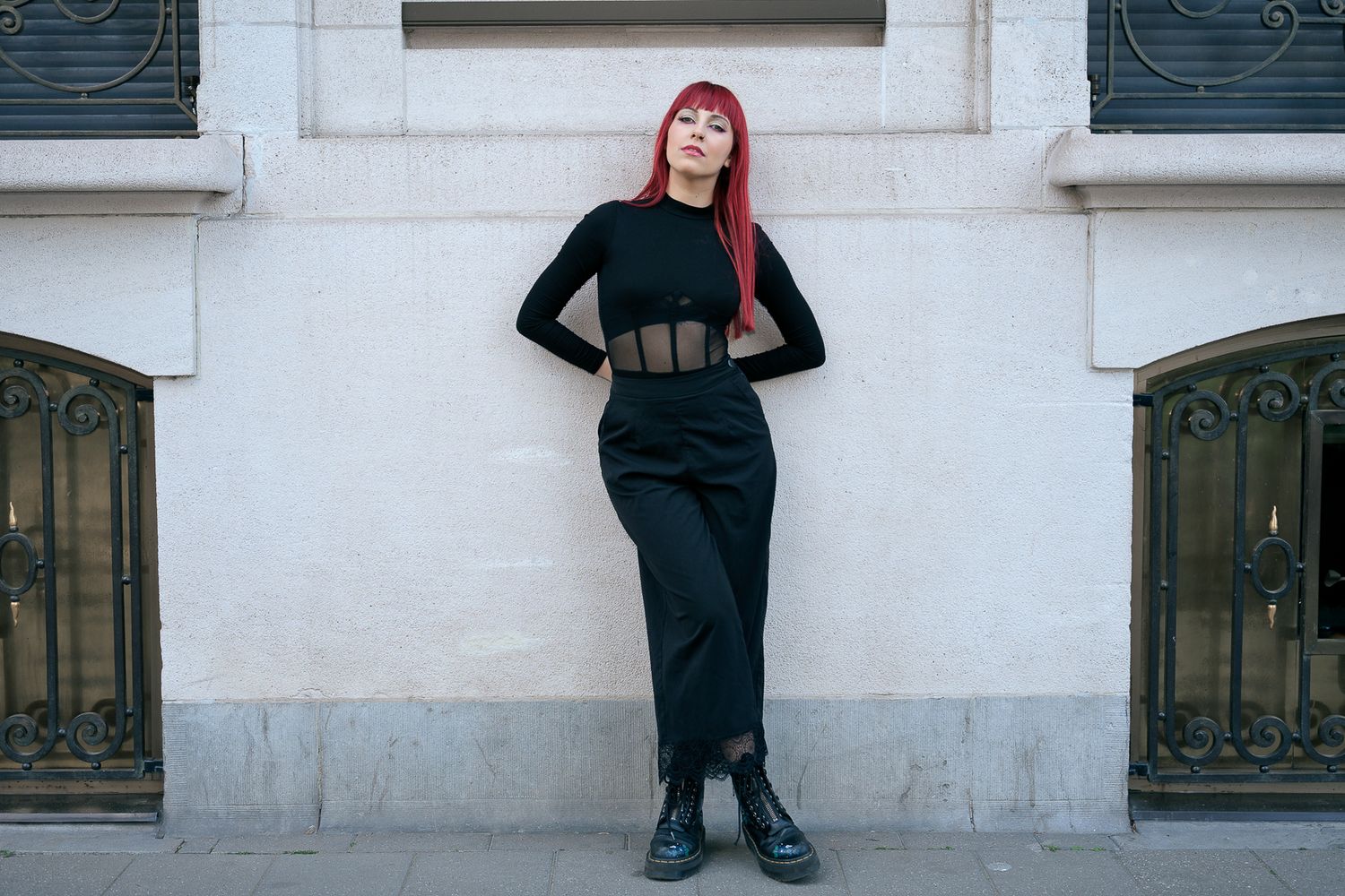 Red-haired woman in black midi skirt and top standing against white building facade.