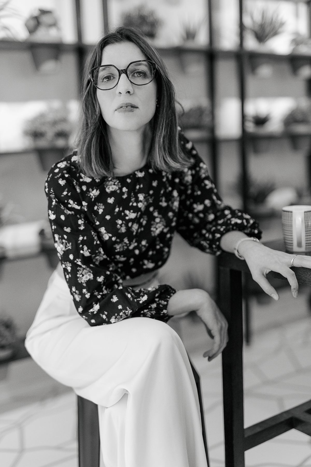 Black and white portrait of a young woman in floral top and wide-leg pants, wearing glasses, seated outdoors.
