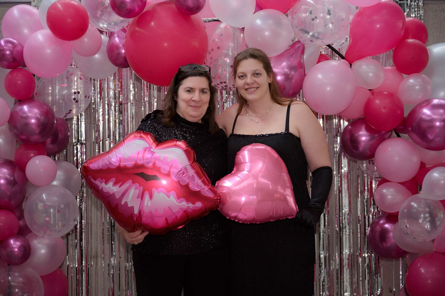 Two women pose with pink heart and lips balloons in front of a pink and white balloon arch backdrop.