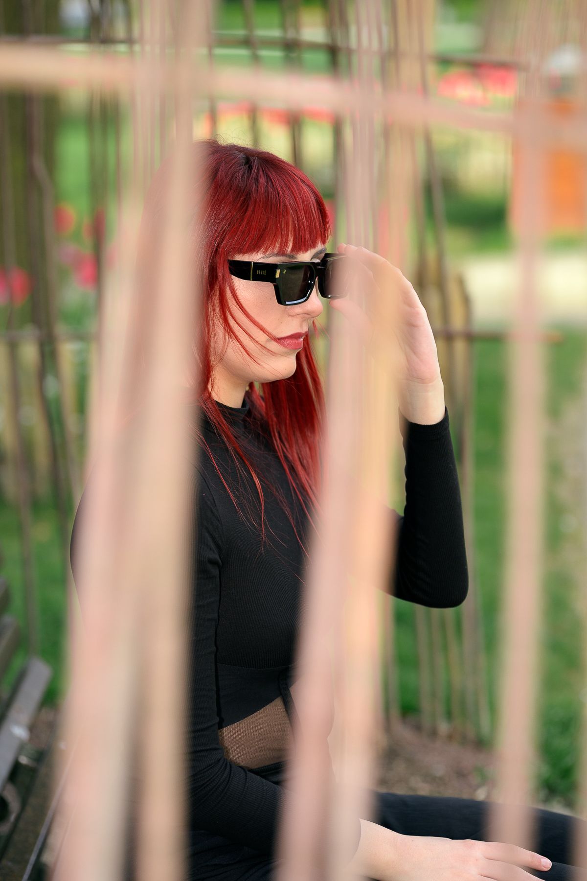 Red-haired woman in black outfit and sunglasses posing outdoors behind wooden slats in a park.