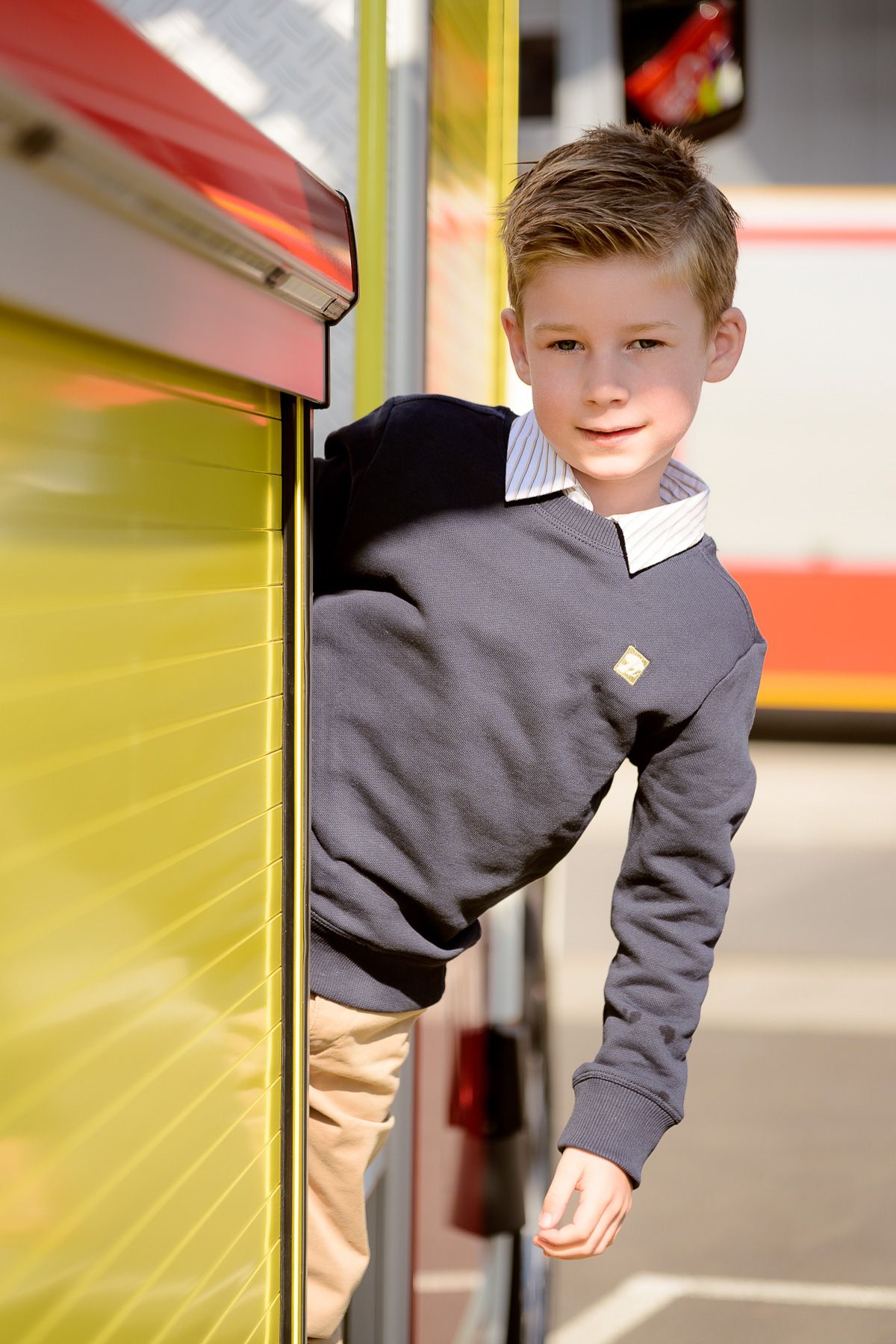 Young boy in gray sweater and khaki pants standing near a yellow and red vehicle door.