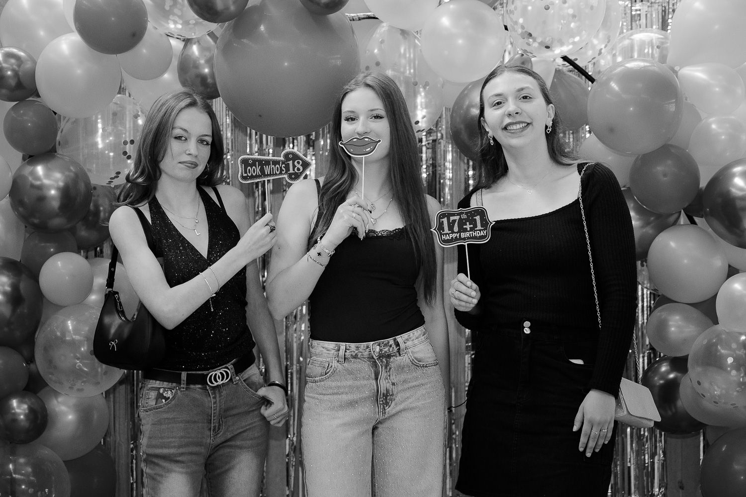 Three young women pose with decorative props in front of a balloon wall at a celebration party.
